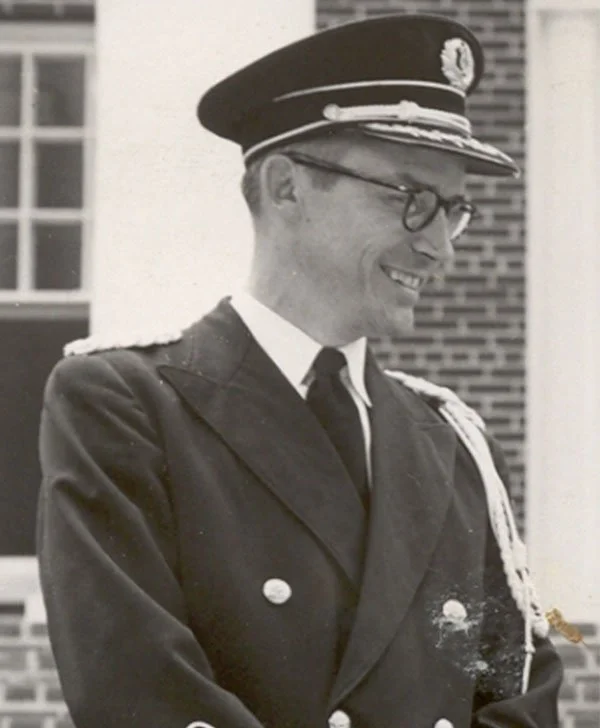 A black and white photo of a smiling man in a formal military uniform, wearing glasses and a peaked cap, standing outdoors in front of a brick building.