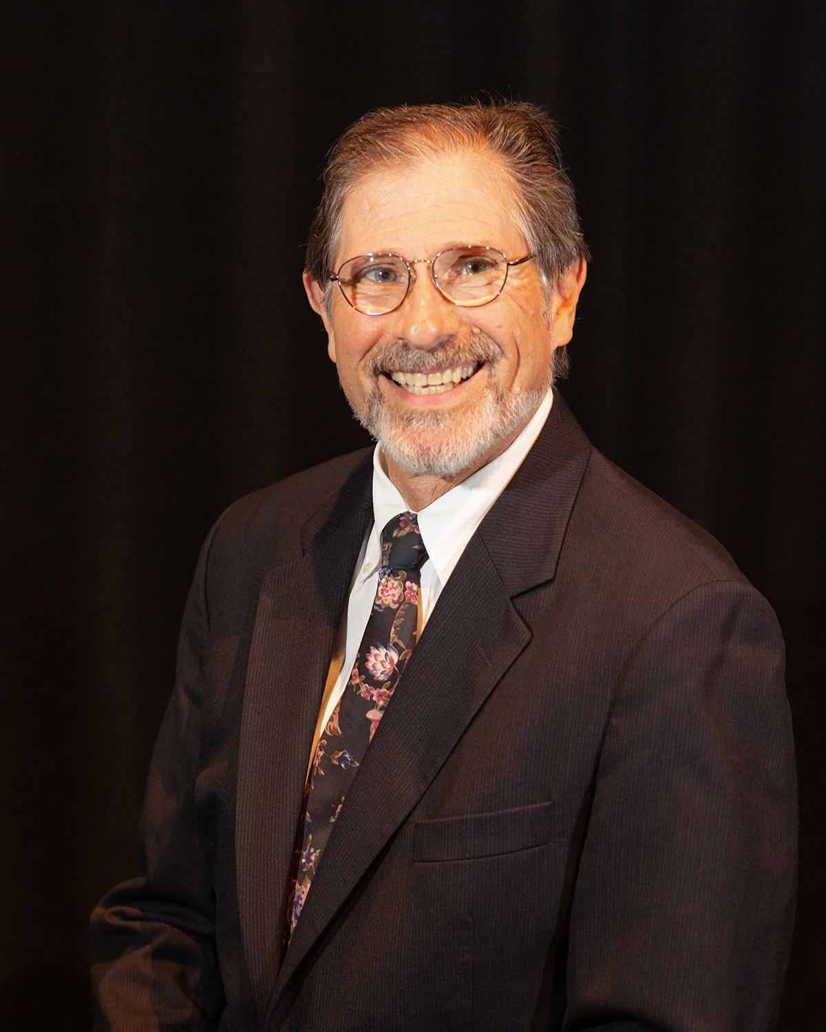 A middle-aged man with glasses, graying hair, and a beard, dressed in a dark suit, white shirt, and floral tie, smiling against a black background.