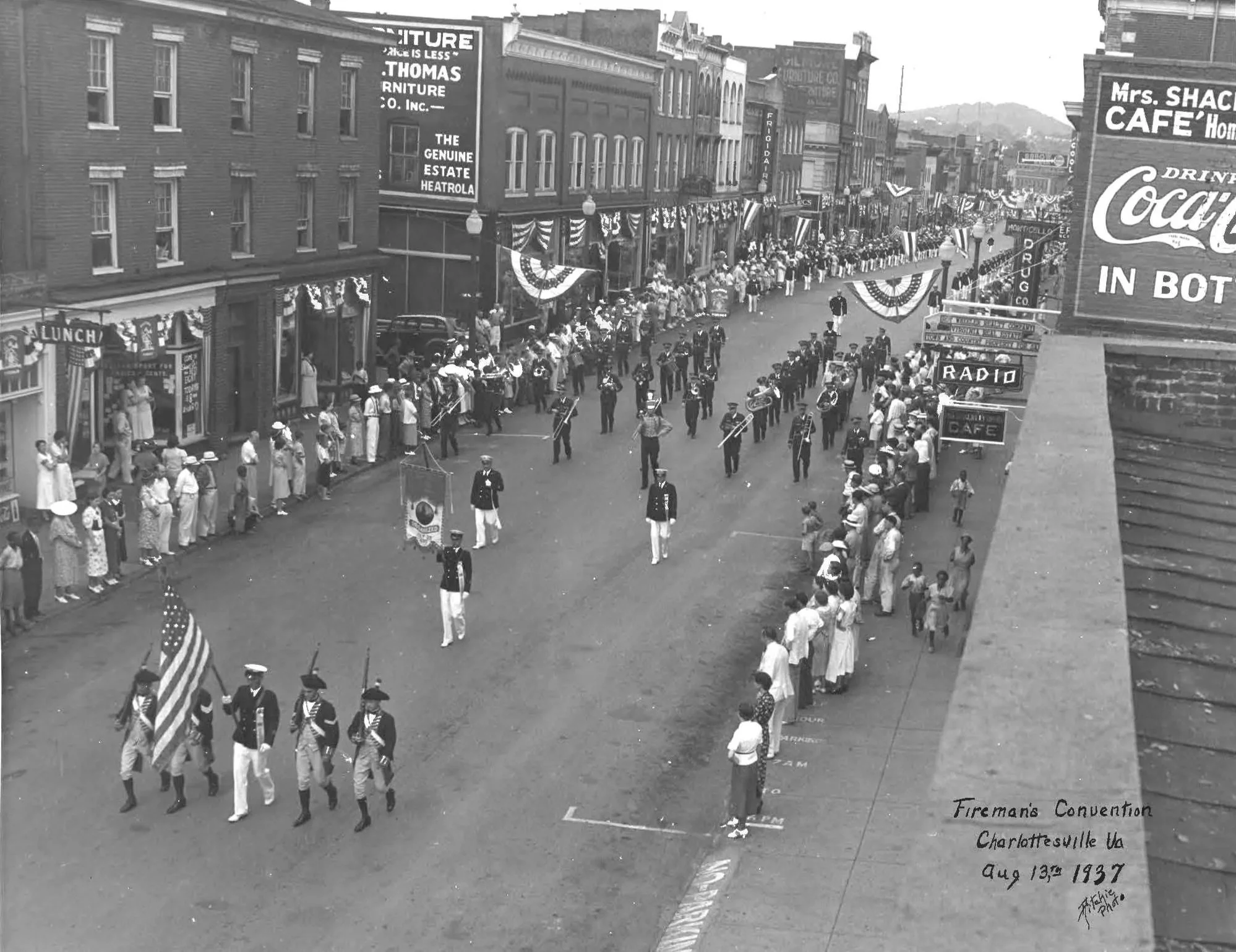 A black and white photo capturing a parade in downtown Charlottesville, Virginia, during the Fireman's Convention on August 13th, 1937. The street is decorated with patriotic bunting, and people are gathered along the sidewalks watching the parade, which includes uniformed marchers and a local band. The buildings along the street display various signs and advertisements, including furniture stores and cafes.