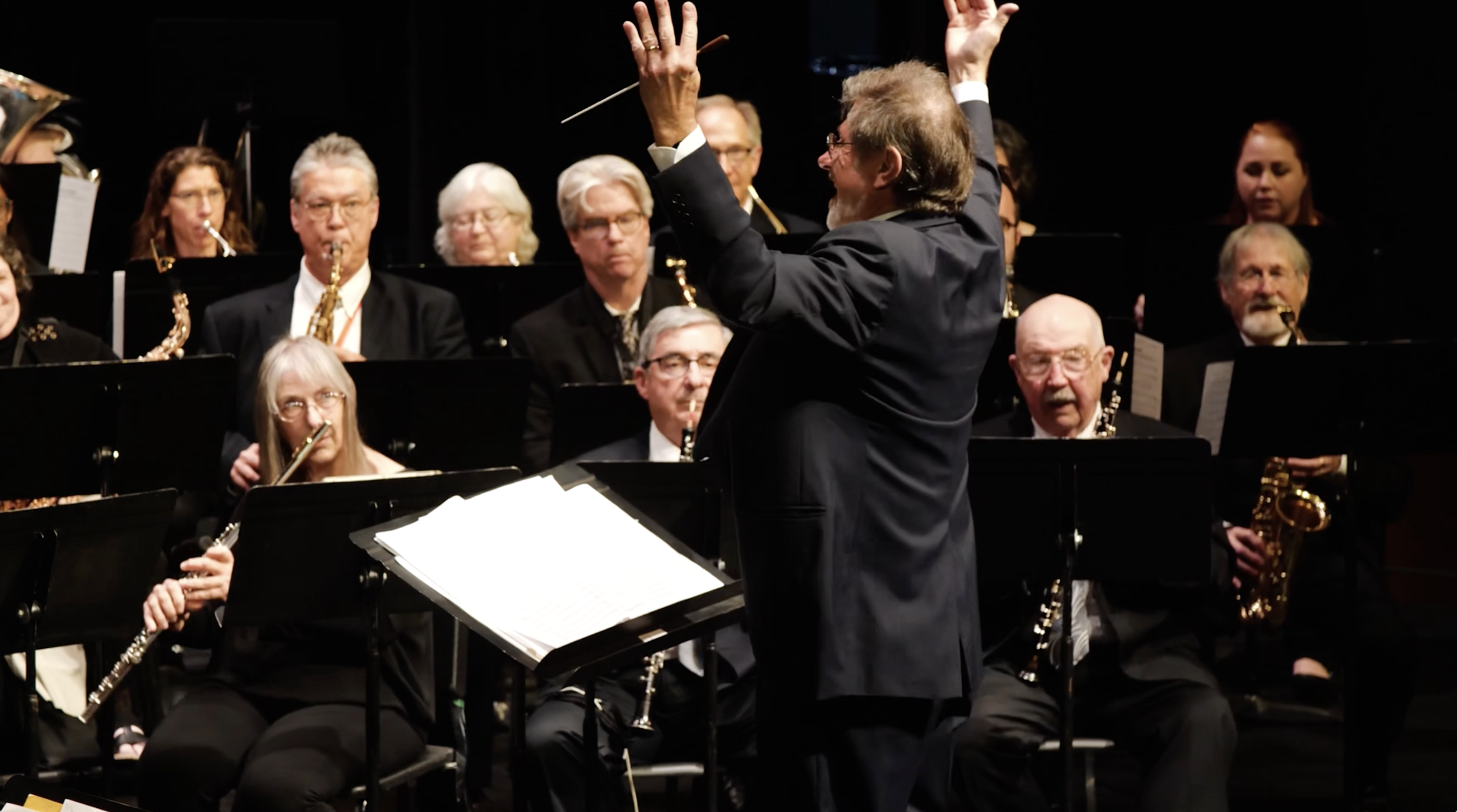 An orchestra performing on stage with a conductor leading them. The musicians are playing wind instruments, and some are reading sheet music. The conductor is standing in front, raising one hand and holding a baton, guiding the performance.
