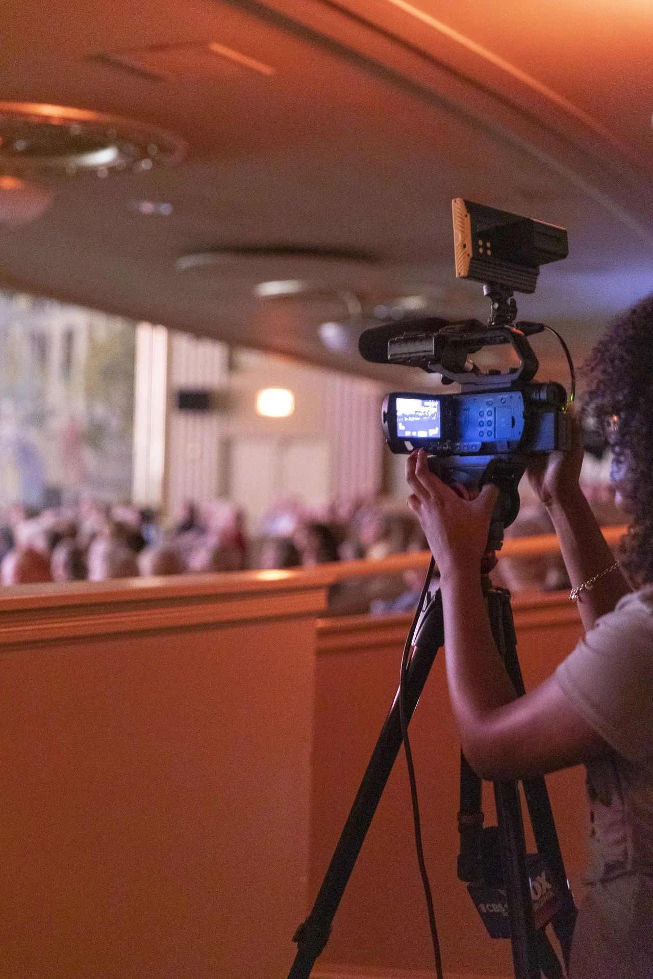 Camera operator filming a performance or event in a dimly lit theater or auditorium with a blurred audience in the background.