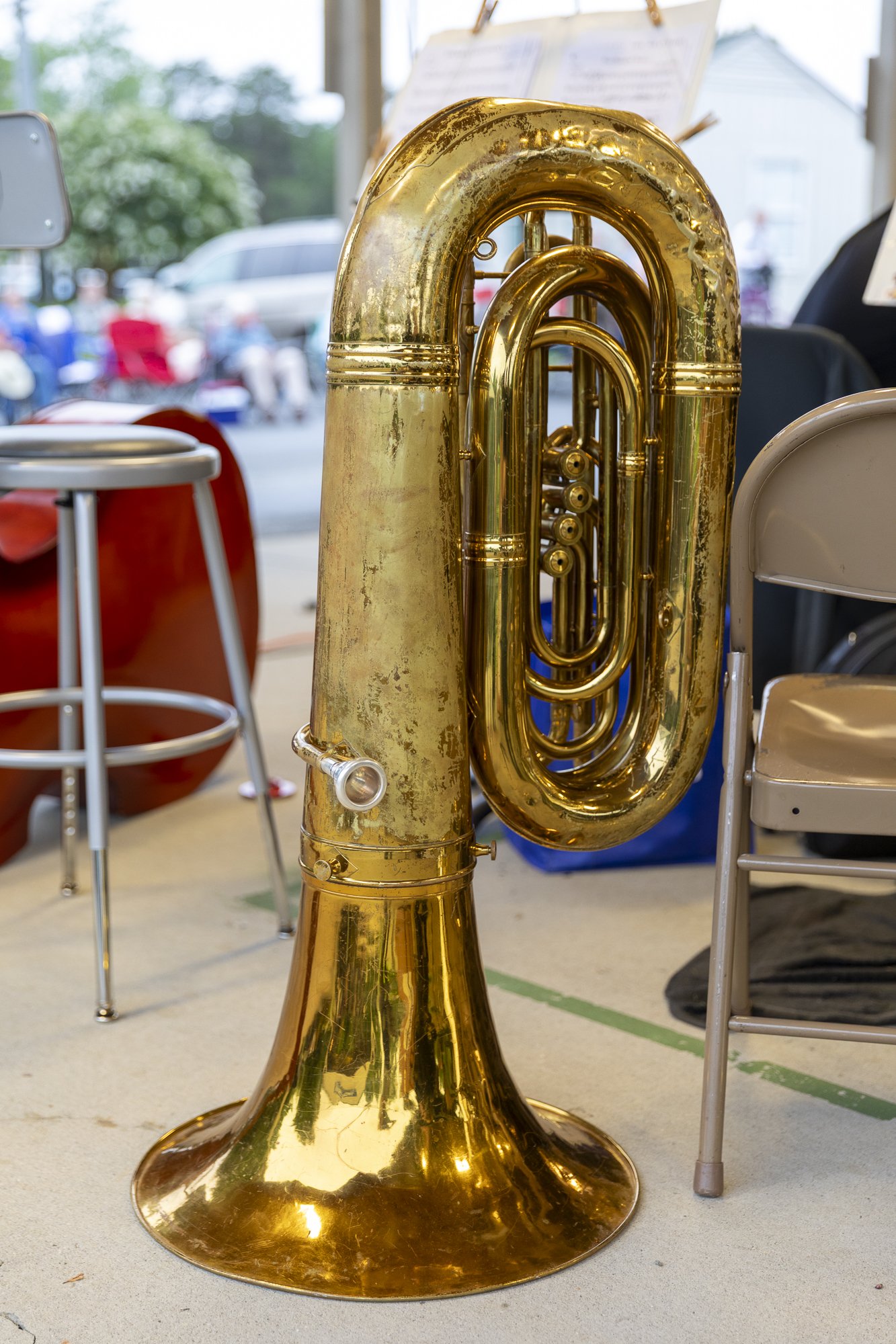 A shiny gold-colored tuba is positioned on the floor among chairs and a small table in a room with large windows, outdoor scenery visible through the windows.