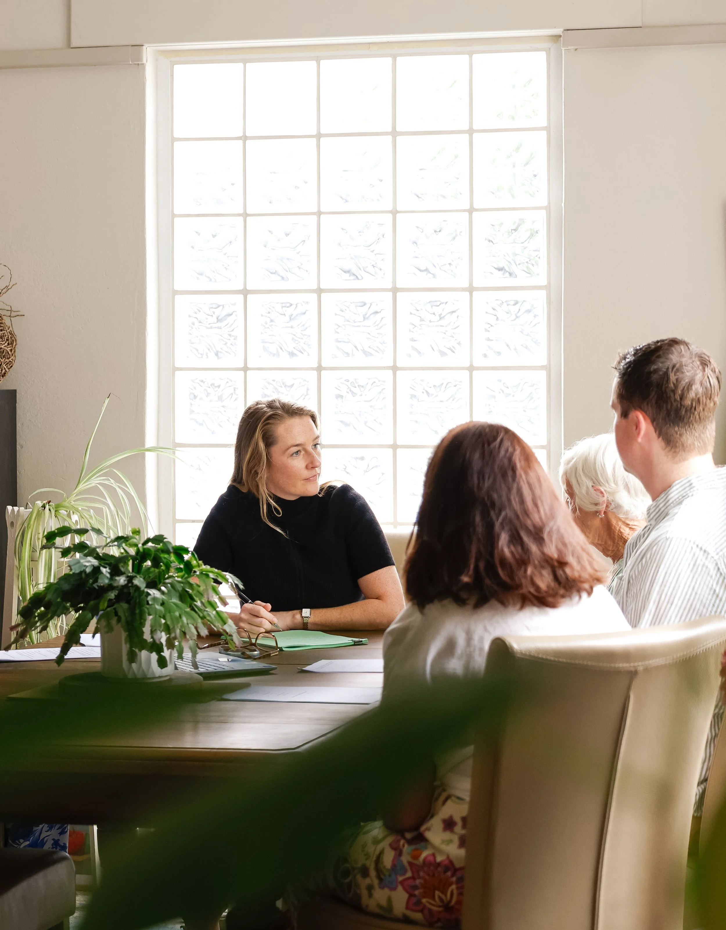 A woman in a black shirt sitting at a table with four other people, engaging in a meeting or discussion, with a large window behind them and a potted plant on the table.