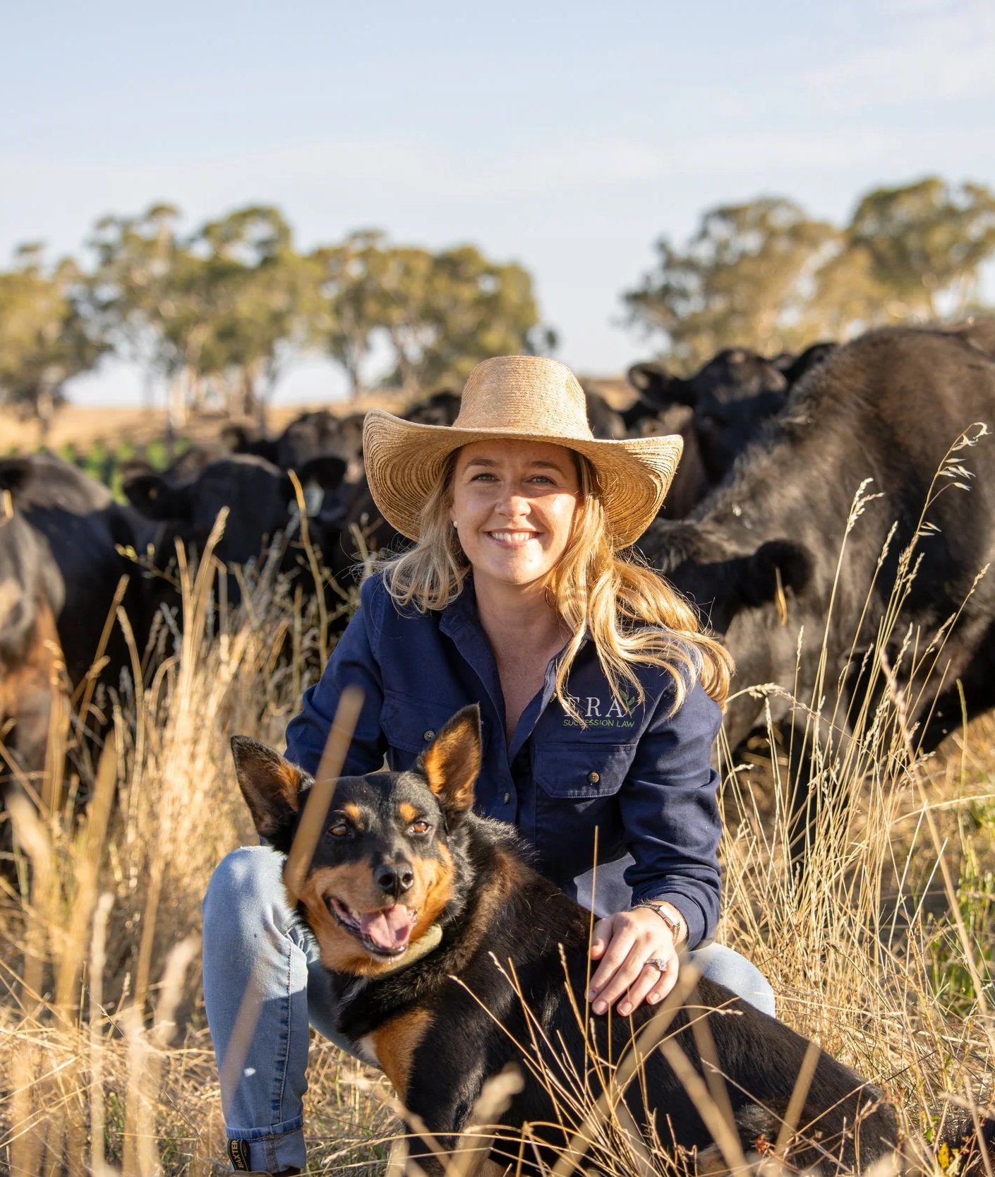 A woman smiling with a cowboy hat and a dog in a field, with cows grazing in the background.