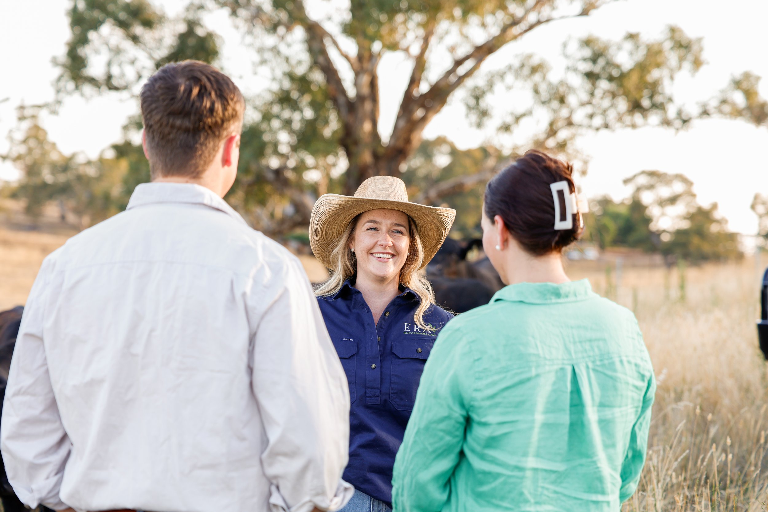 Three people having a conversation outdoors on a farm, with cows and trees in the background. One woman with a wide-brimmed hat is smiling while talking to a man and a woman, seen from the back.