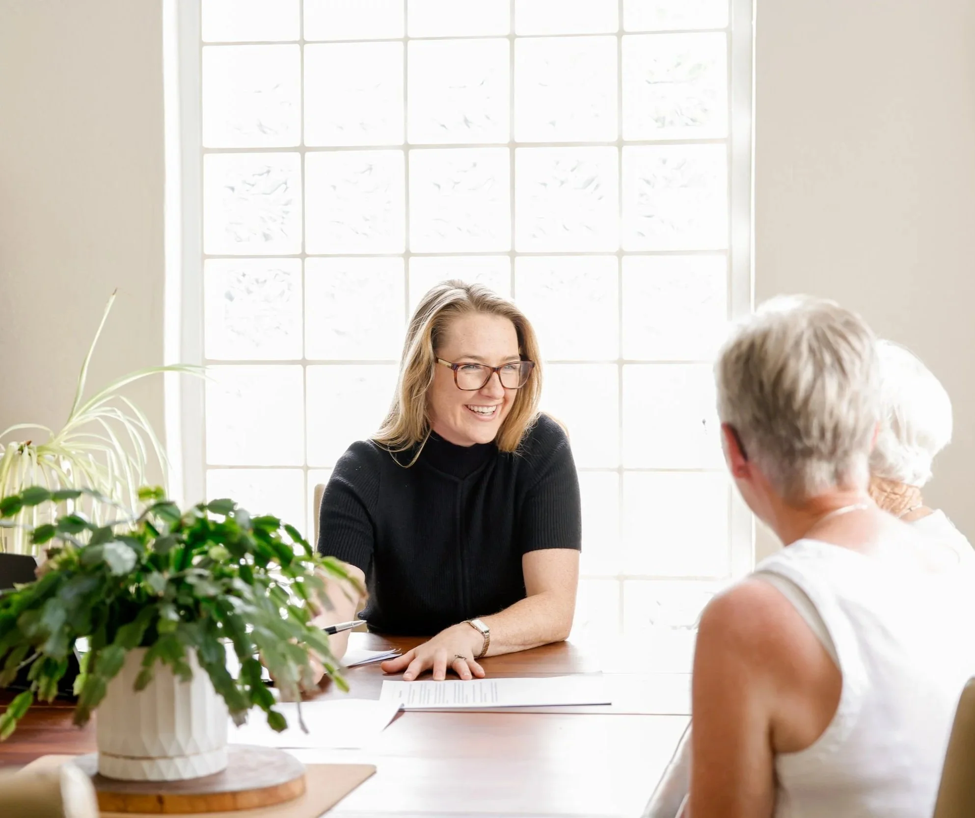 Three women having a conversation in a bright room, with a woman in black seated at a table with green plants and papers, smiling and talking to two other women with gray hair.