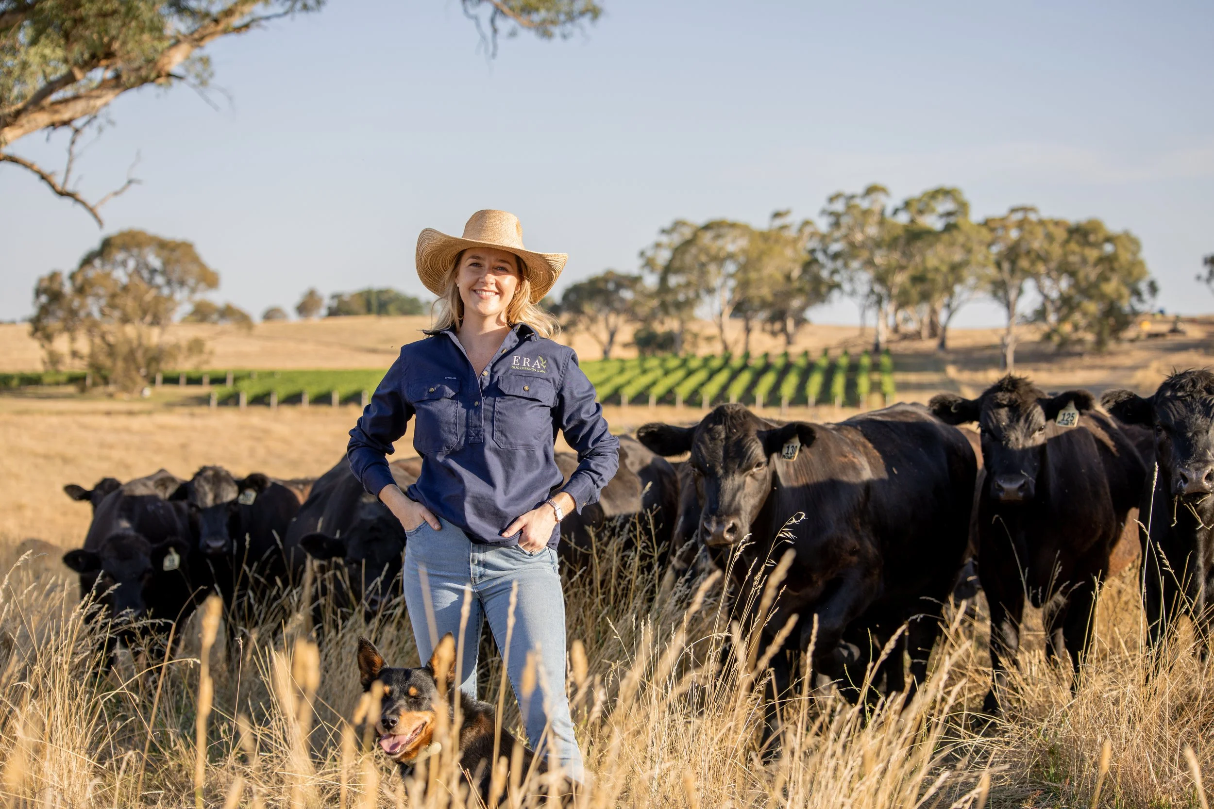 A woman standing in a field with tall grass, wearing a large sun hat and a navy blue shirt, smiling, with a black dog in front of her, and a herd of black calves behind her. There are trees and cultivated land in the background on a sunny day.