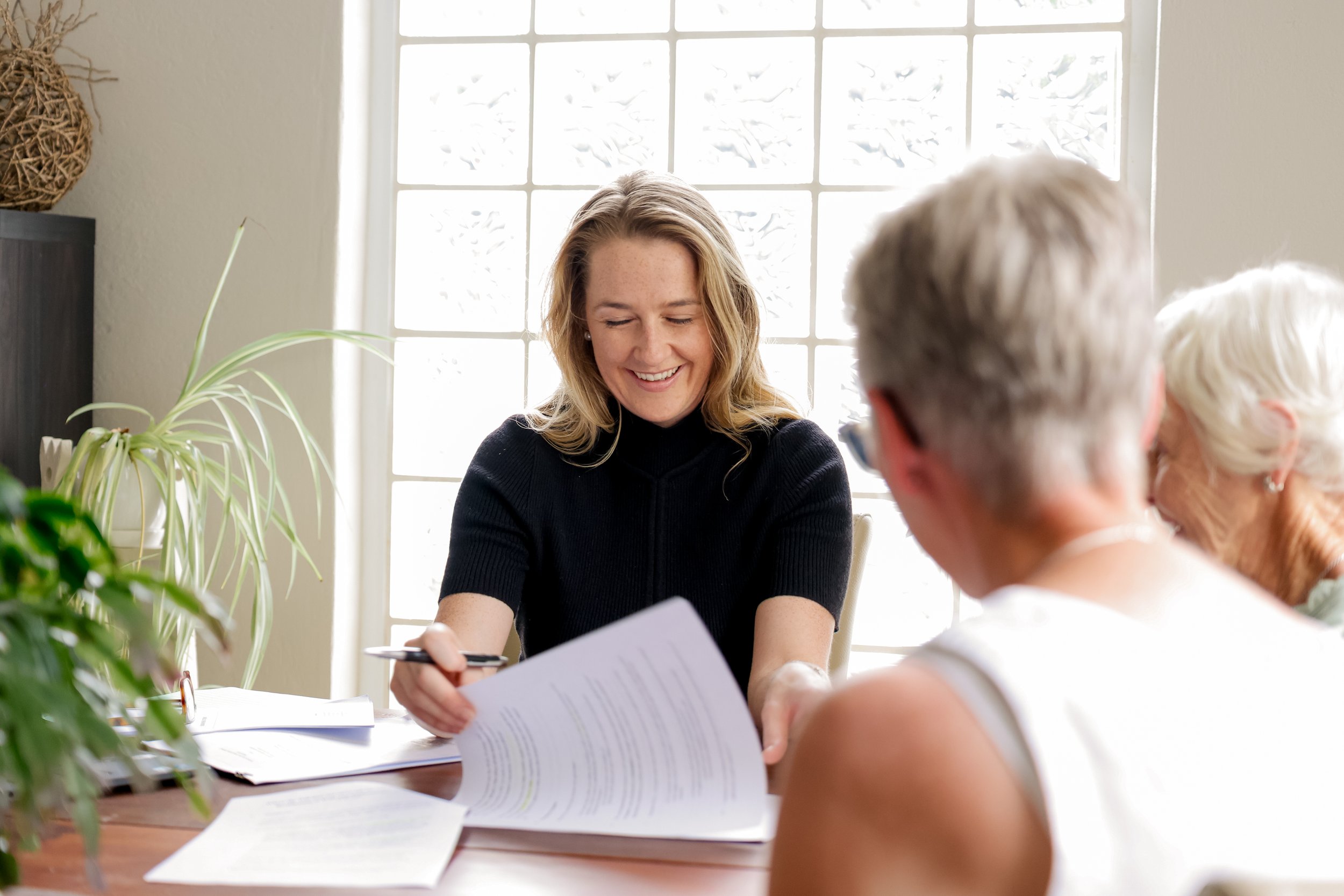 A woman smiling and holding a pen, showing a document to two elderly women sitting at a table with papers, in a bright room with large window.