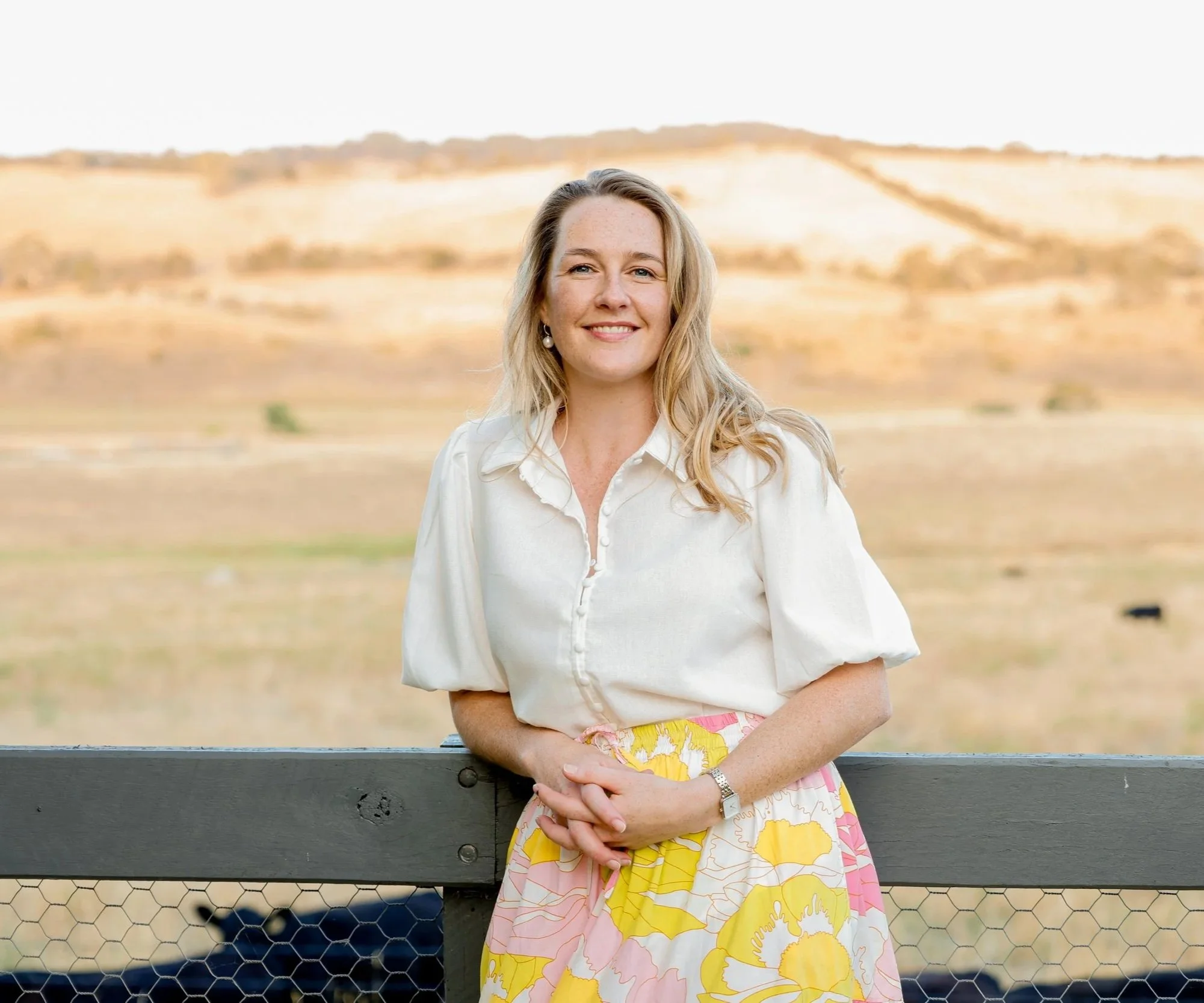 A woman with blonde hair wearing a white blouse and a colorful floral skirt, standing outdoors near a fence with a scenic hilly landscape in the background.