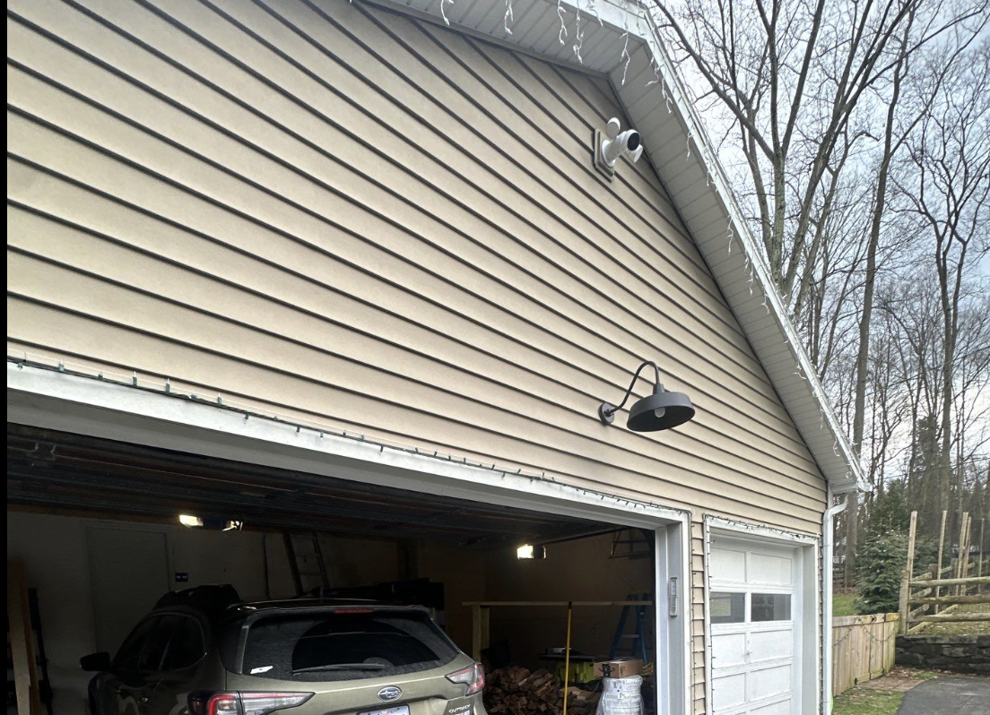 Outside view of a beige garage with a partially open door, a black lamp on the wall, and Christmas lights on the roof edge. Inside the garage, a car is parked along with wood and other stored items. The garage is attached to a house with trees visible in the background.