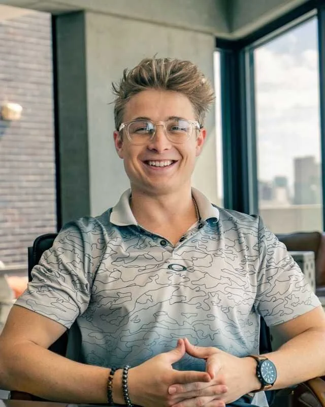 Smiling young man with glasses, wearing a patterned polo shirt, sitting indoors near large windows with a view of city buildings