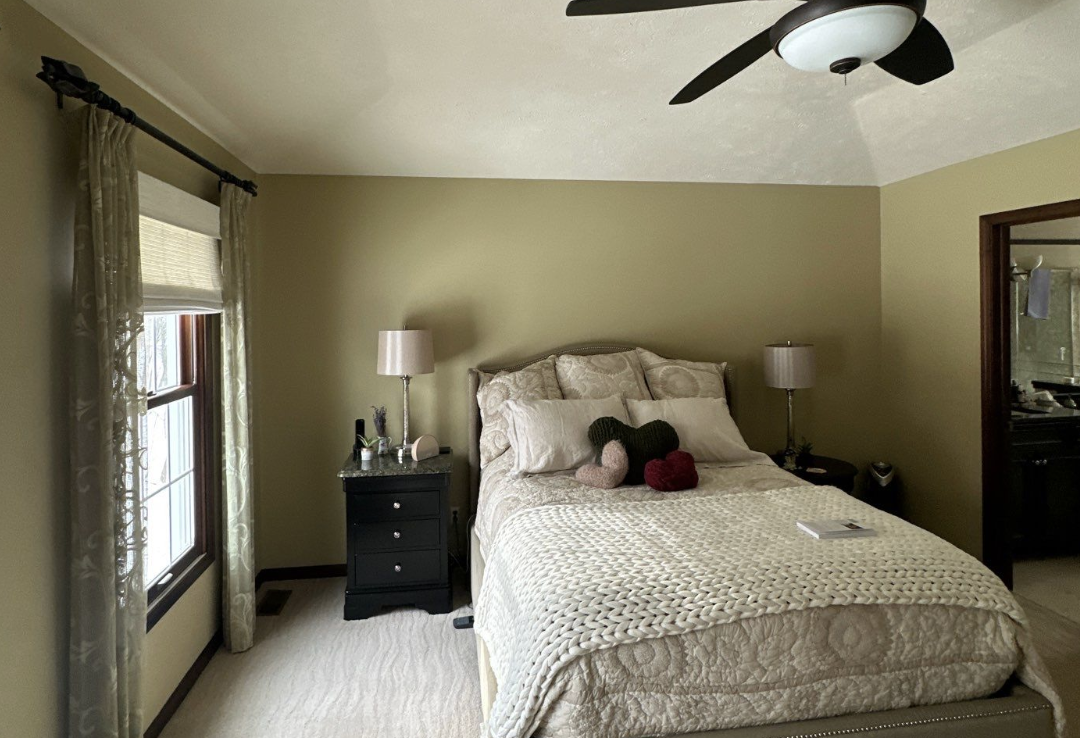 A bedroom with a bed, bedside tables with lamps, and a ceiling fan. The bed has multiple pillows and plush toys. There's a window with curtains on the left side.