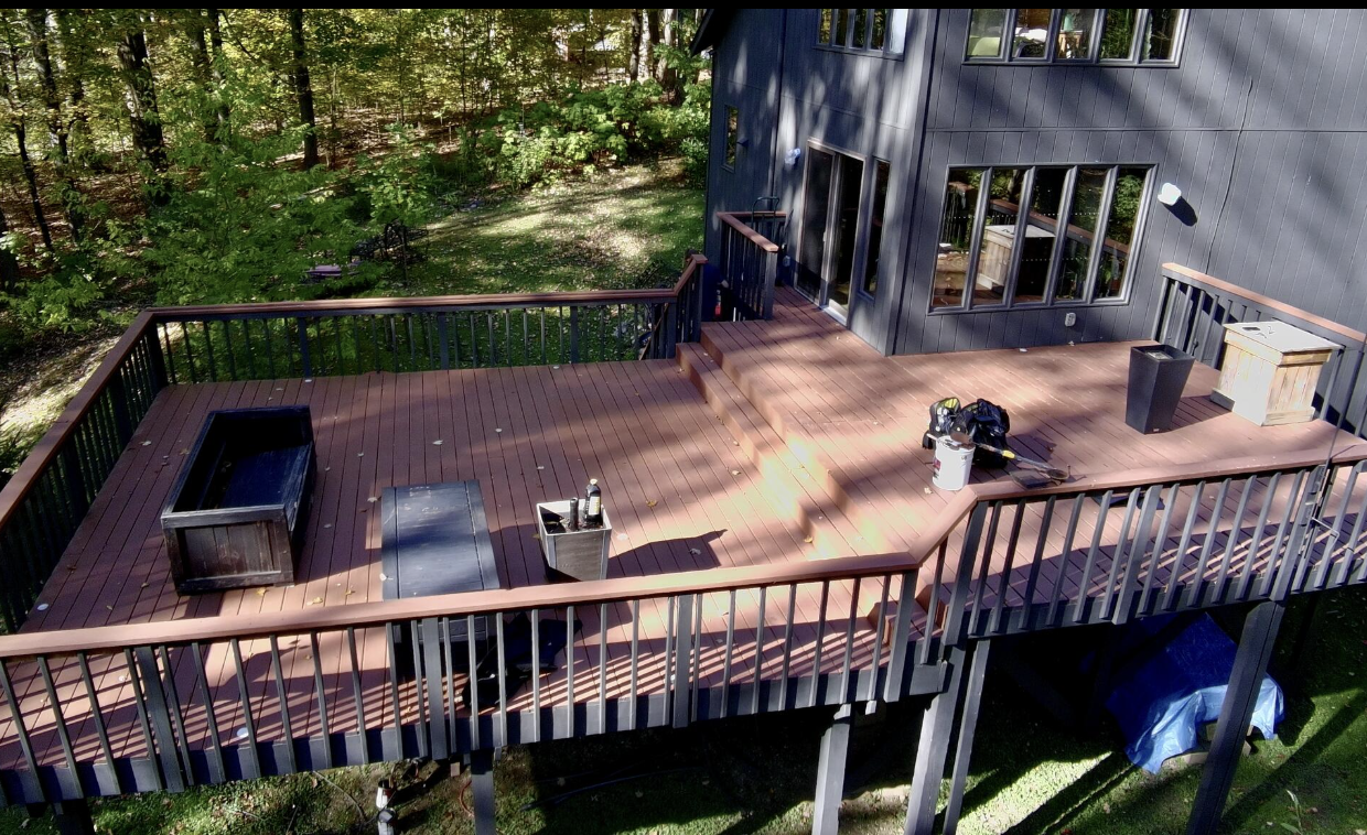 An elevated wooden deck attached to a house, with tools and storage containers on it, overlooking a green wooded backyard.