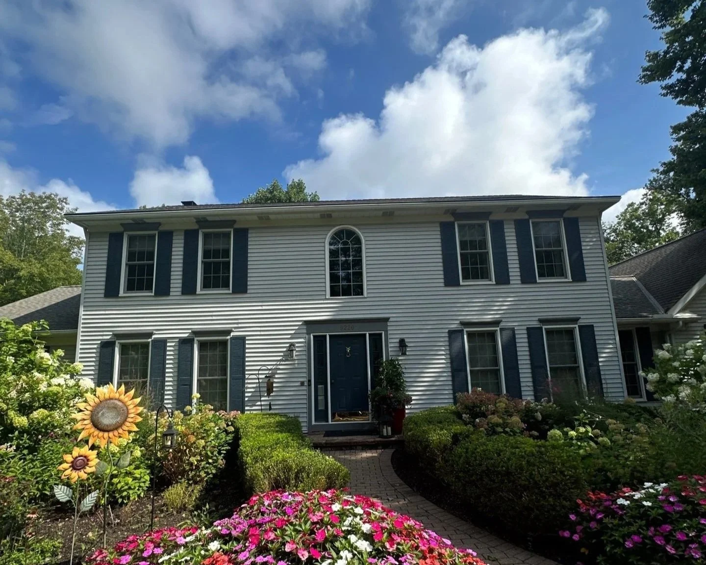 Front view of a white two-story house with dark blue shutters, surrounded by colorful flowers and green bushes under a partly cloudy sky.