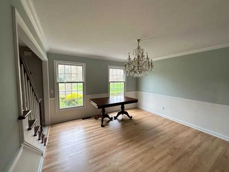 Empty dining room with two large windows, a wooden table, a chandelier, hardwood floors, and light-colored walls with white trim.