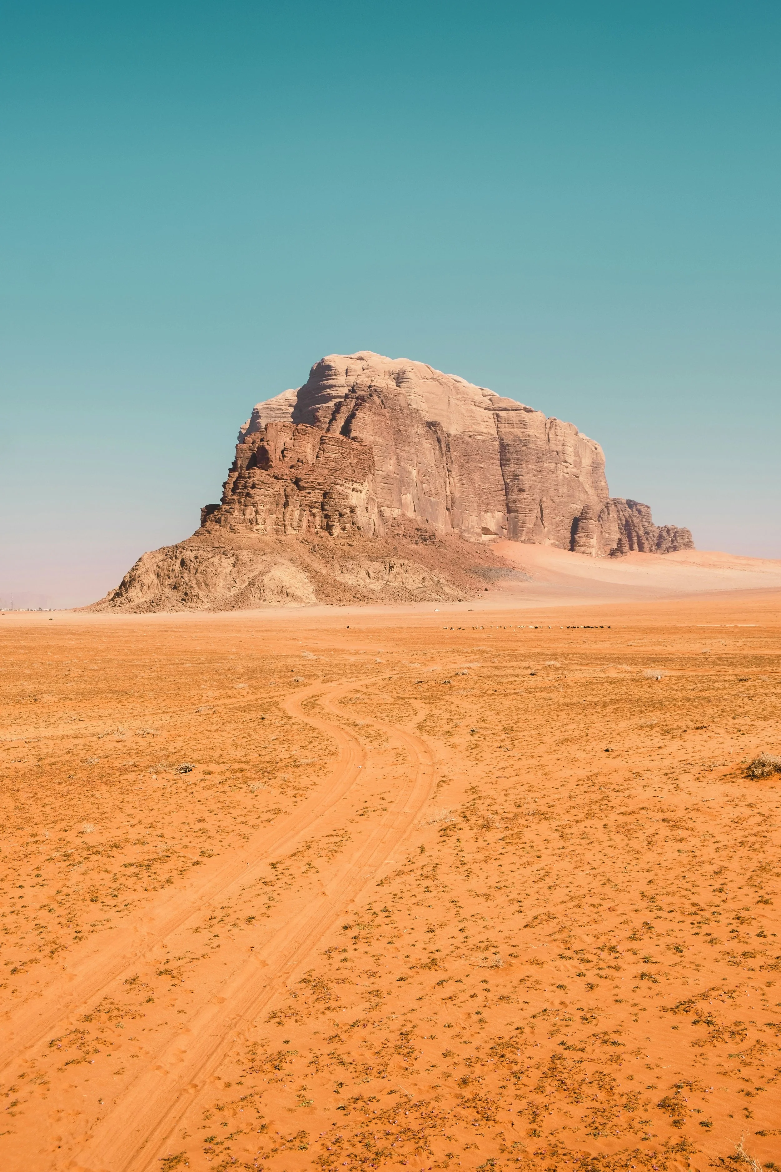 A large sandstone formation in a desert landscape with a dirt track leading toward it and a clear blue sky above.