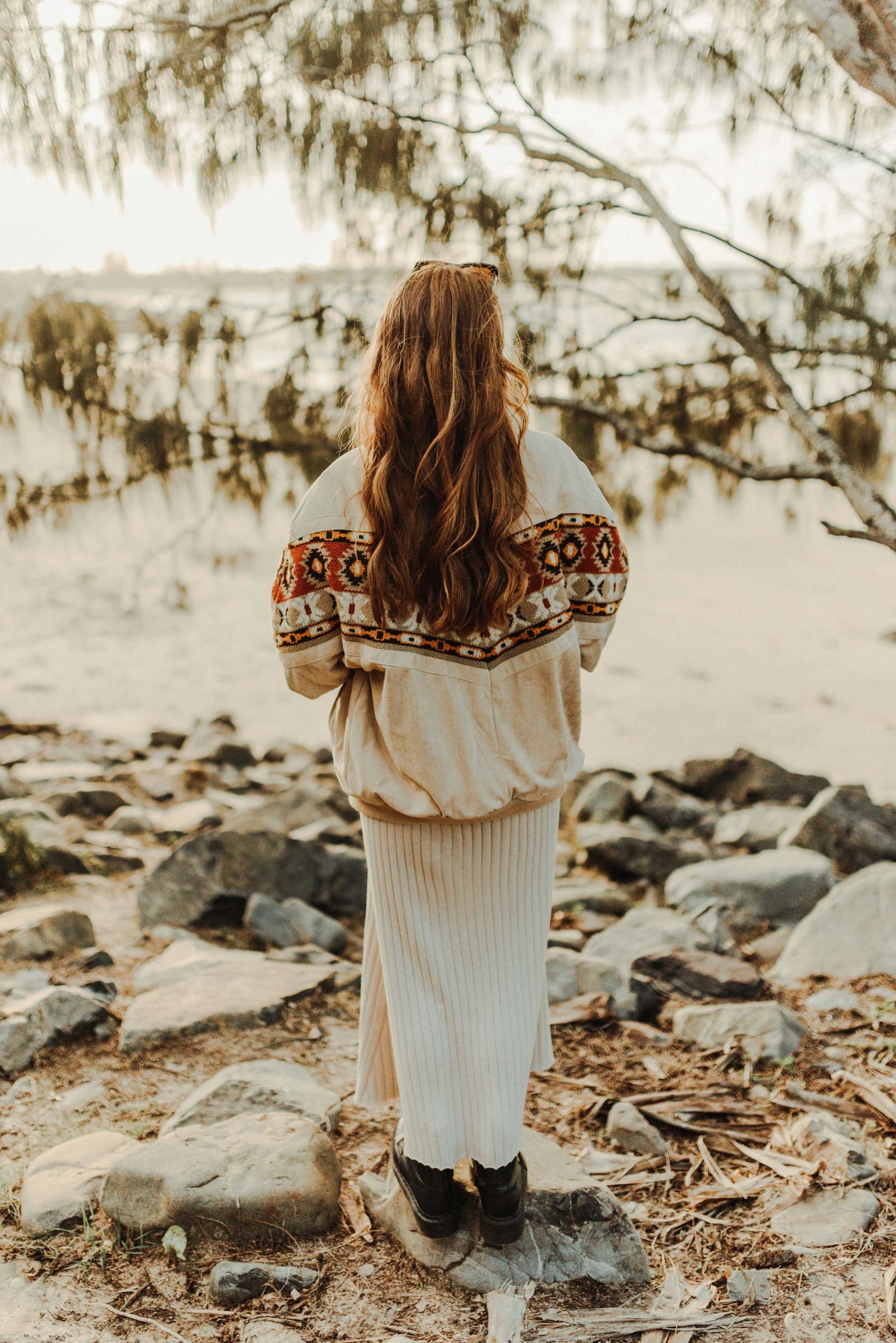 A woman with long, wavy brown hair standing on rocks near a body of water, facing away, during sunset.