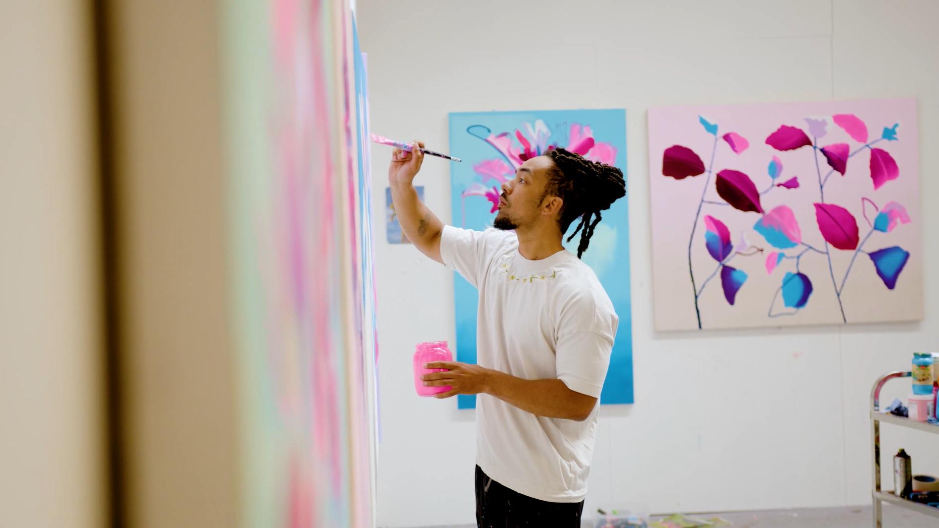 A male artist with dreadlocks painting on a wall in an art studio, holding a pink paint jar in one hand and a paintbrush in the other, with colorful floral paintings on the wall behind him.