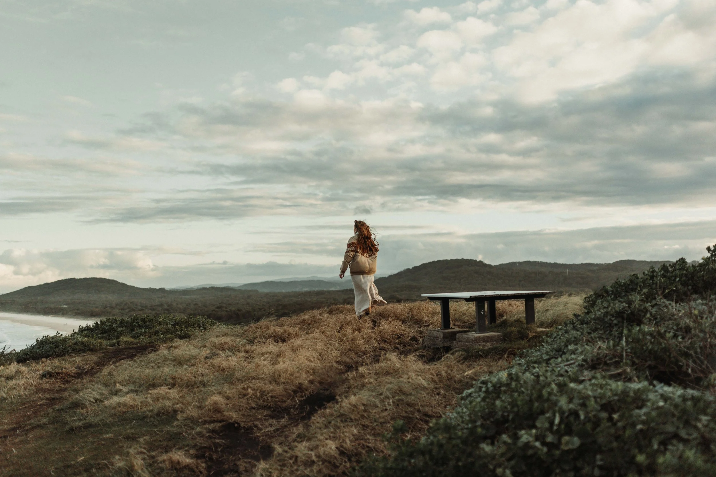 A woman with long red hair walking on a grassy hilltop near the coast during cloudy weather.