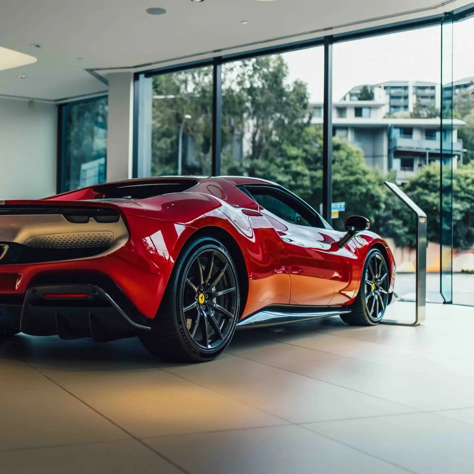 Red sports car inside a showroom with large glass windows showing trees and buildings outside.
