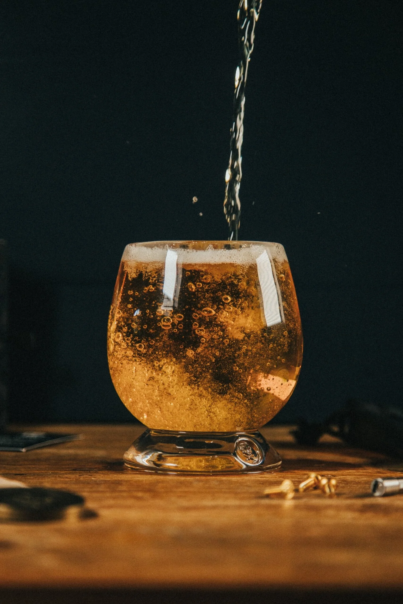 A glass with a short stem being filled with a fizzy, amber-colored beverage, with bubbles rising to the top, placed on a wooden surface against a dark background.