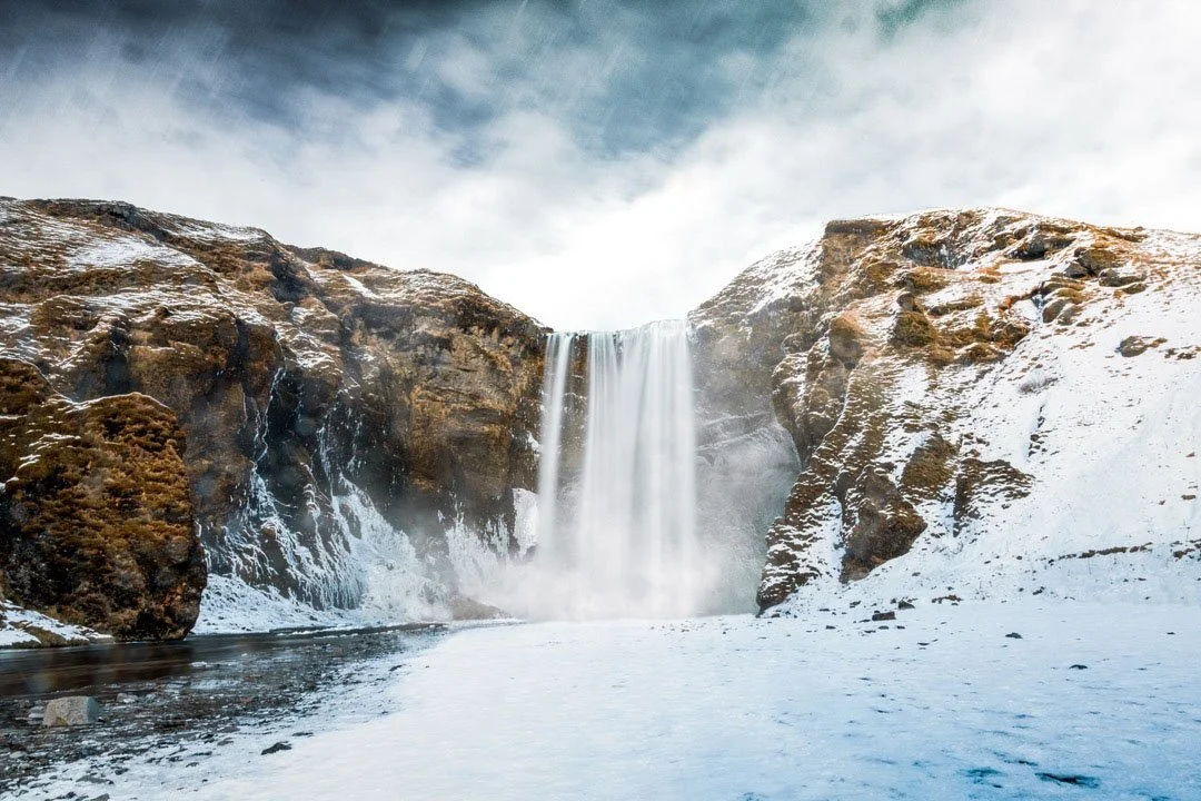 A waterfall in a snowy landscape with snow-covered rocks and cloudy sky