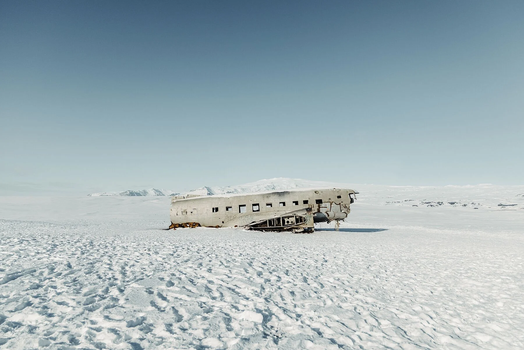 A wrecked airplane lying on snow-covered flat terrain with mountains in the background under a clear blue sky.