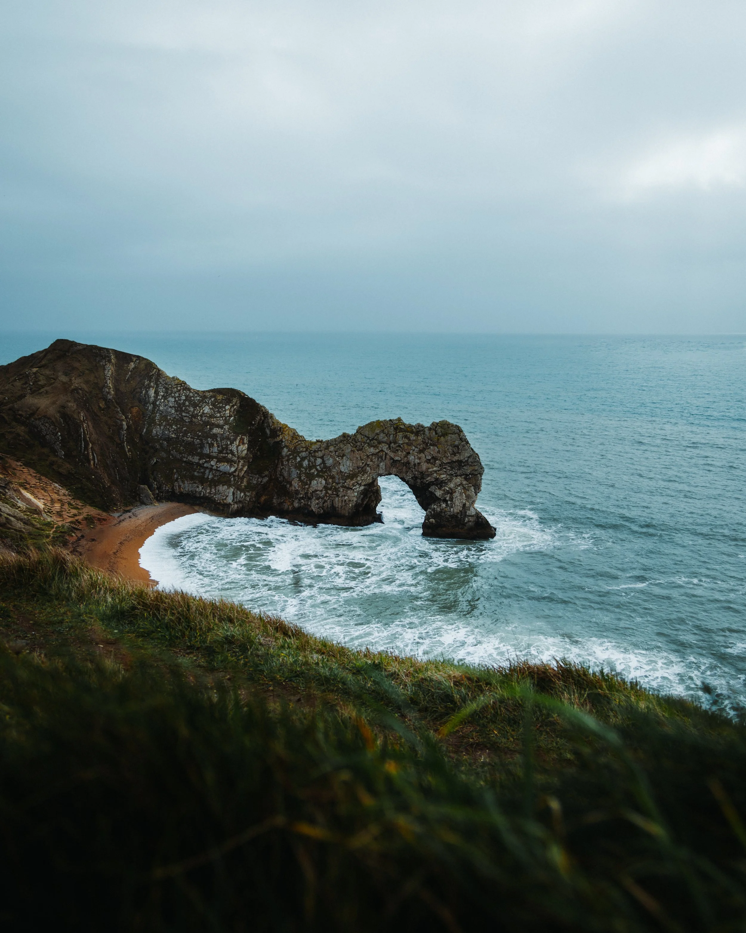 Cliffside with a natural rock arch over the ocean, with grassy foreground and cloudy sky