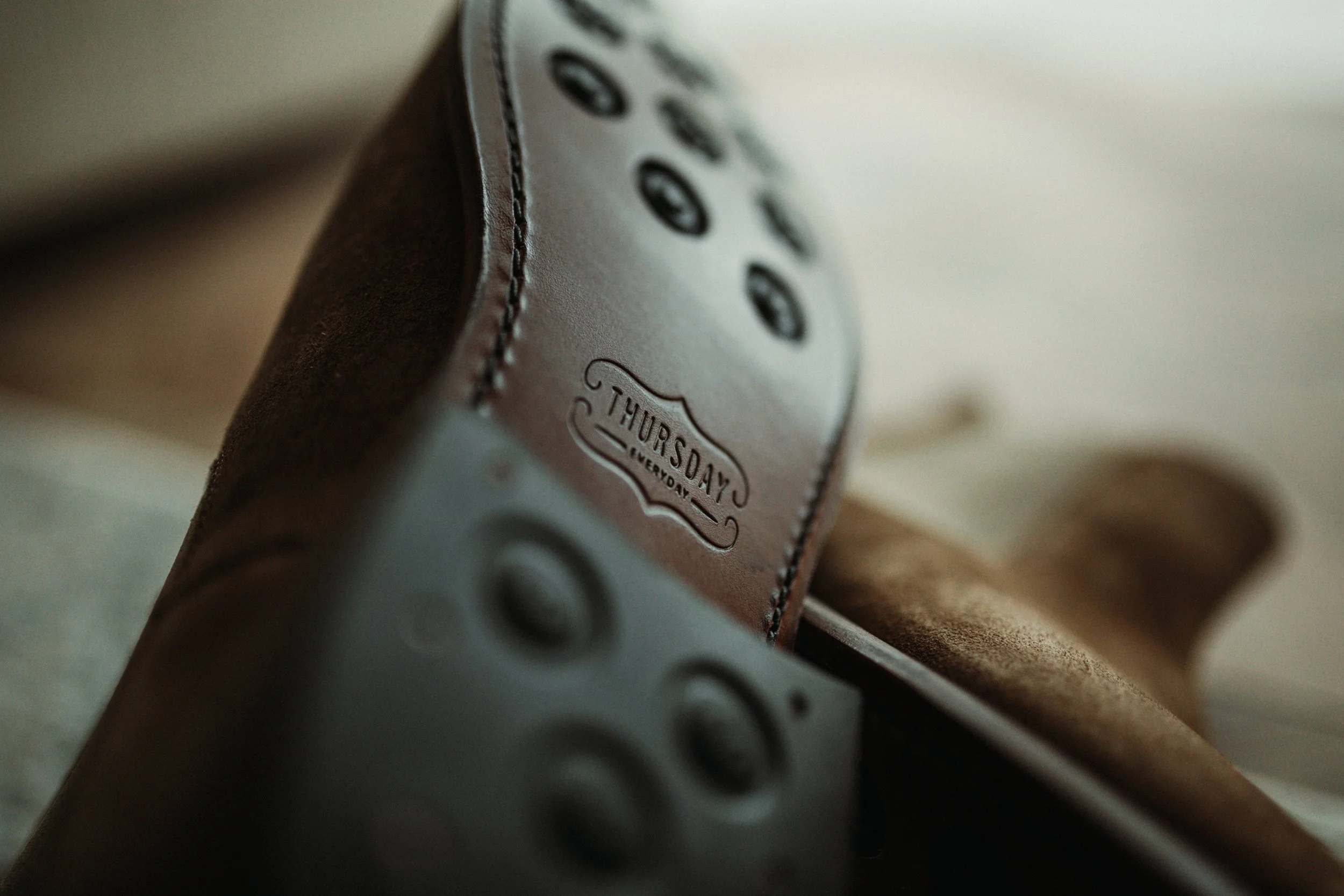 Close-up of a leather strap on a shoe with a stitched mark that reads 'Thursday'. It is attached to a shoe sole with circular nails.