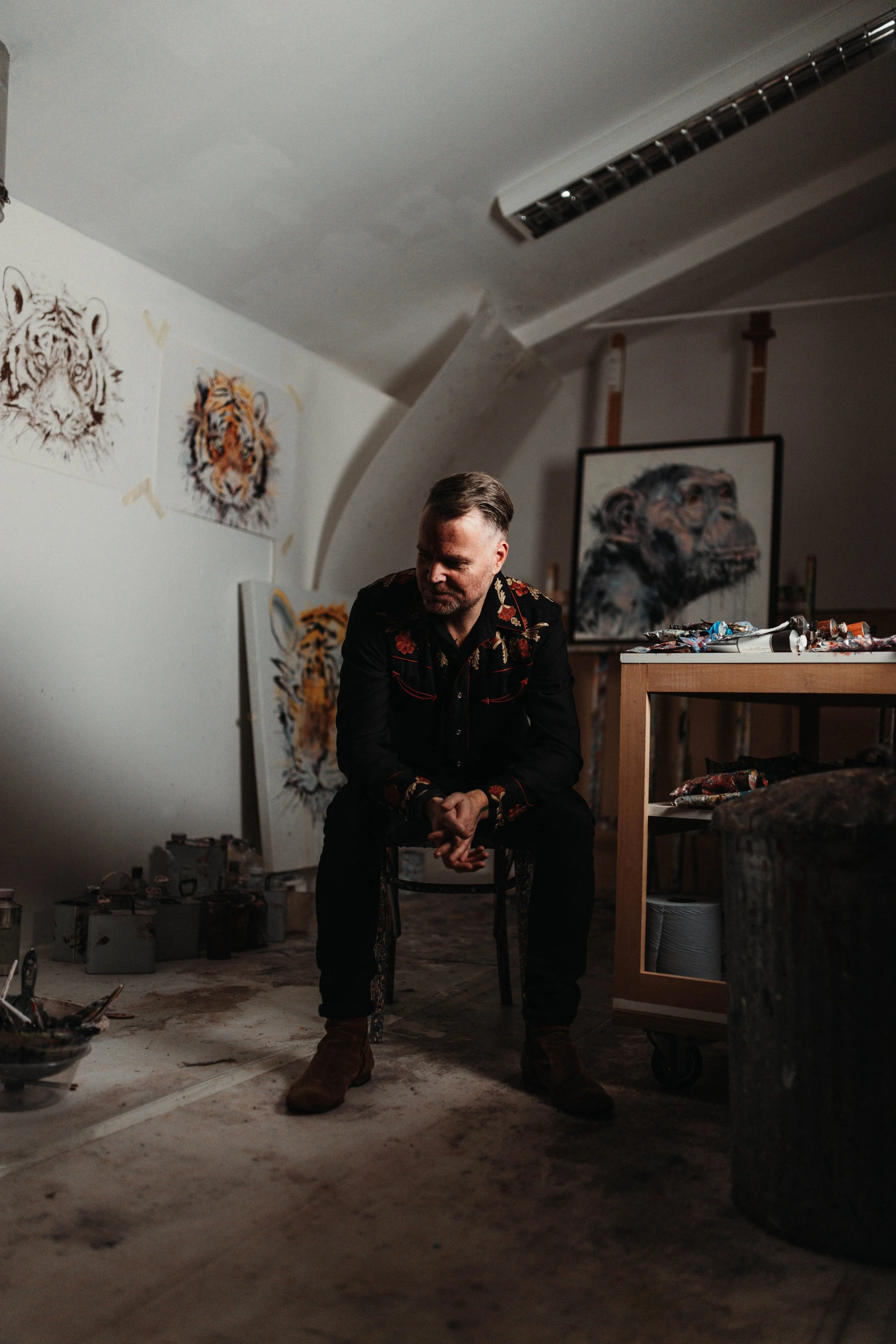 An artist sits on a stool in his studio, surrounded by paintings of tigers and a portrait of a lion. The studio has an arched ceiling, and art supplies and paintings are scattered around.