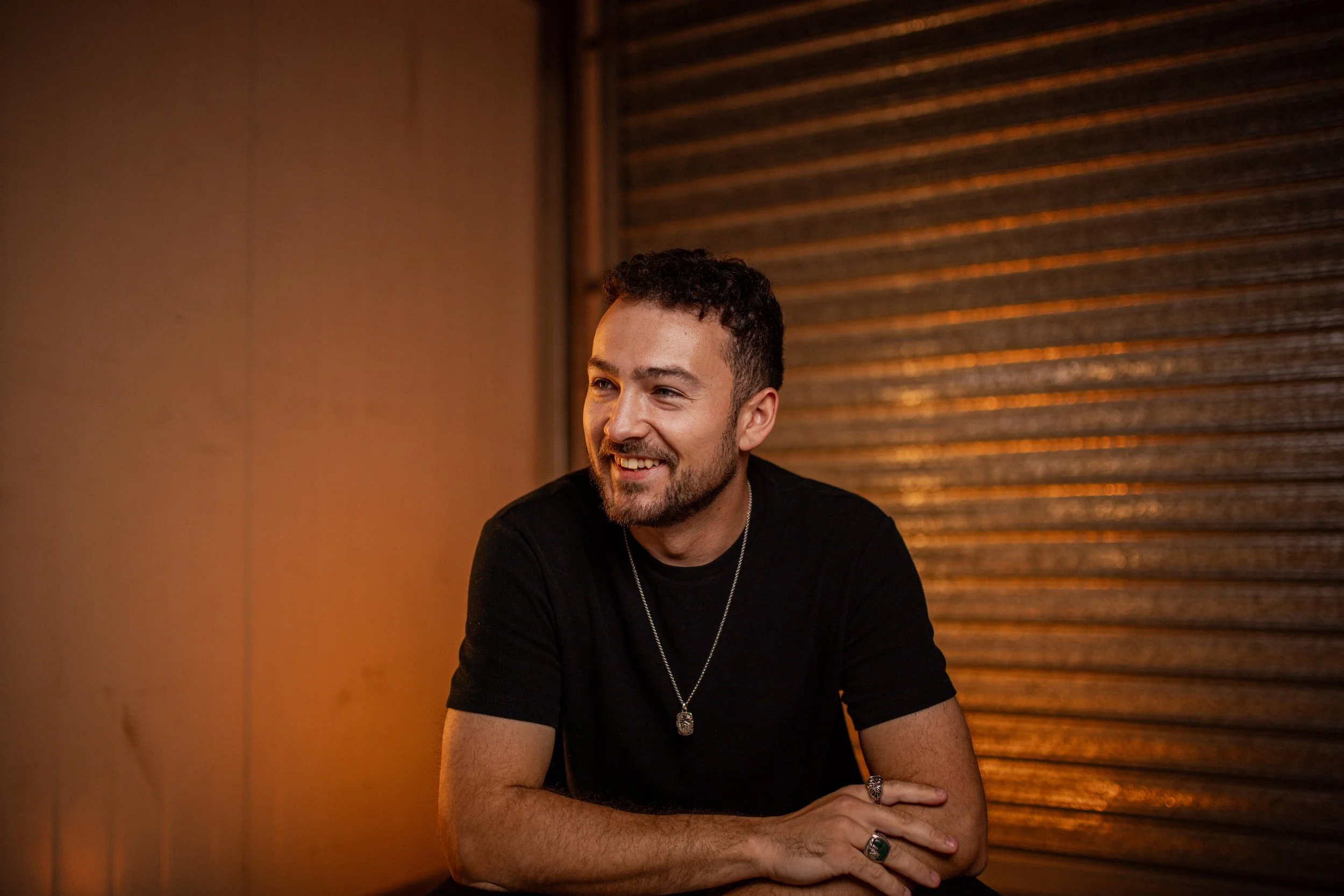 A smiling man with a beard and short curly hair wearing a black t-shirt and silver jewelry, sitting indoors with warm lighting and a wooden wall in background.
