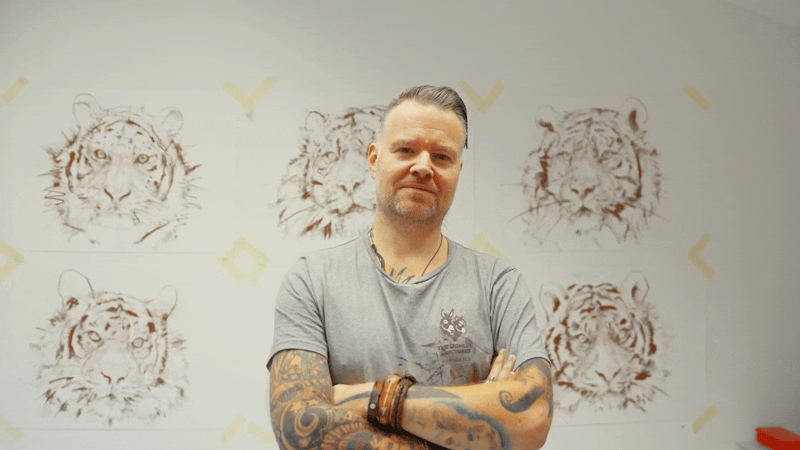 A man with tattoos on his arms stands with his arms crossed in front of a wall with drawings of tiger heads.