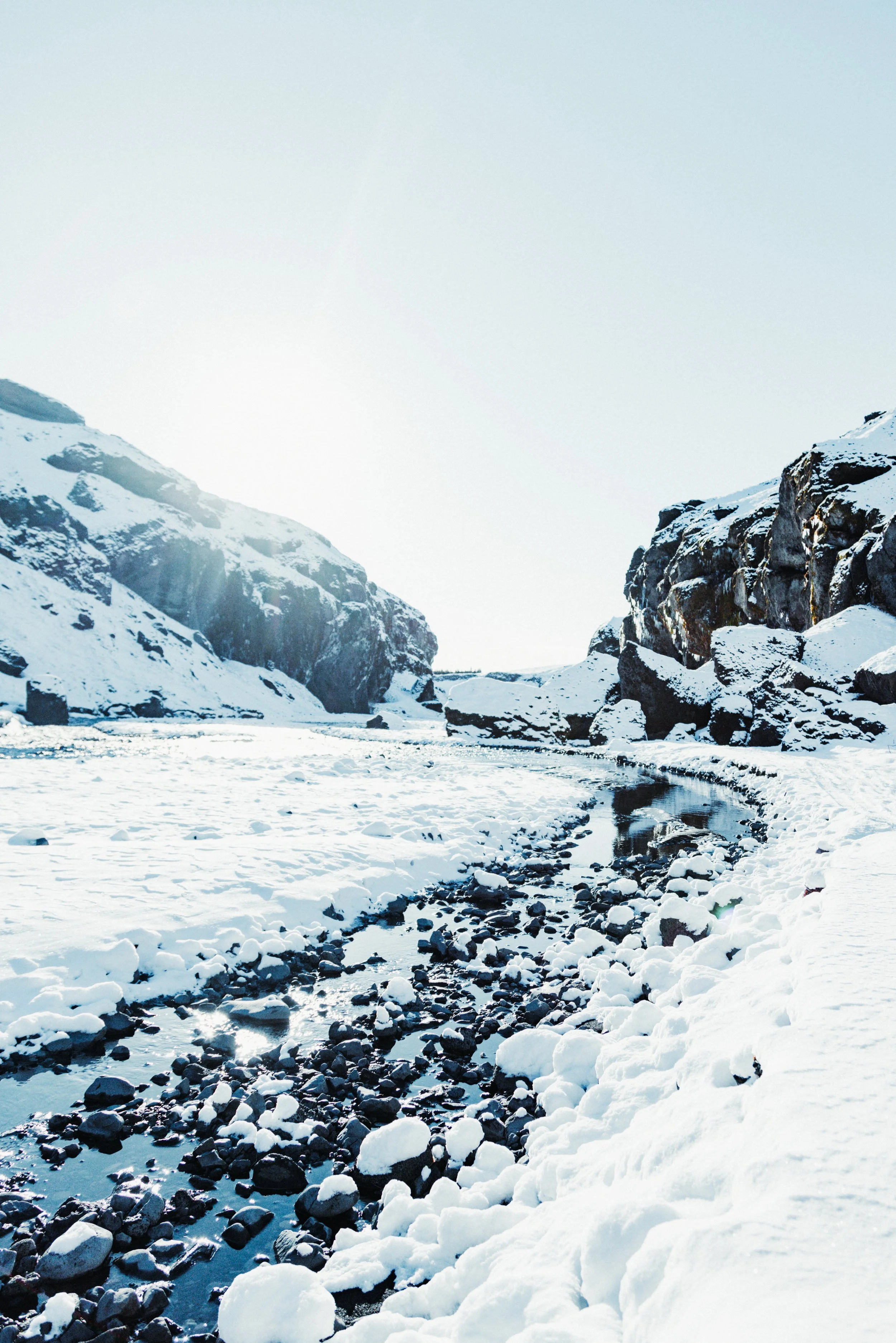 Snow-covered canyon with a narrow river flowing through rocks and cliffs under a bright, cloudy sky.