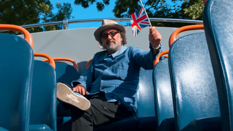 A man with a beard and sunglasses sitting on a bus or tram, holding a small Union Jack flag, wearing a gray hat and blue shirt, with concert seats around him and a background of trees.