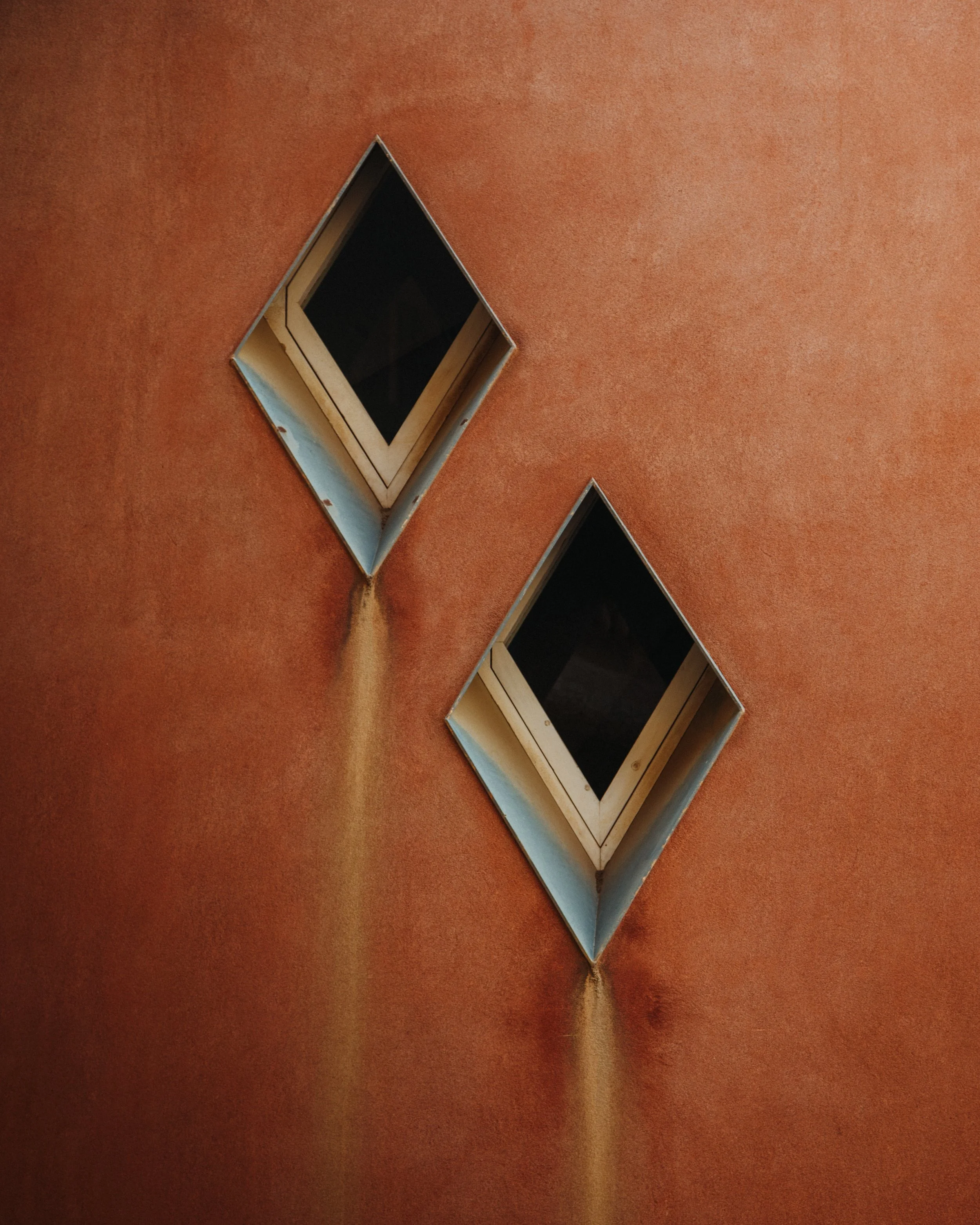Two diamond-shaped attic vents on a rust-colored metal roof, with a stream of rust-colored dust or powder falling from the left vent.