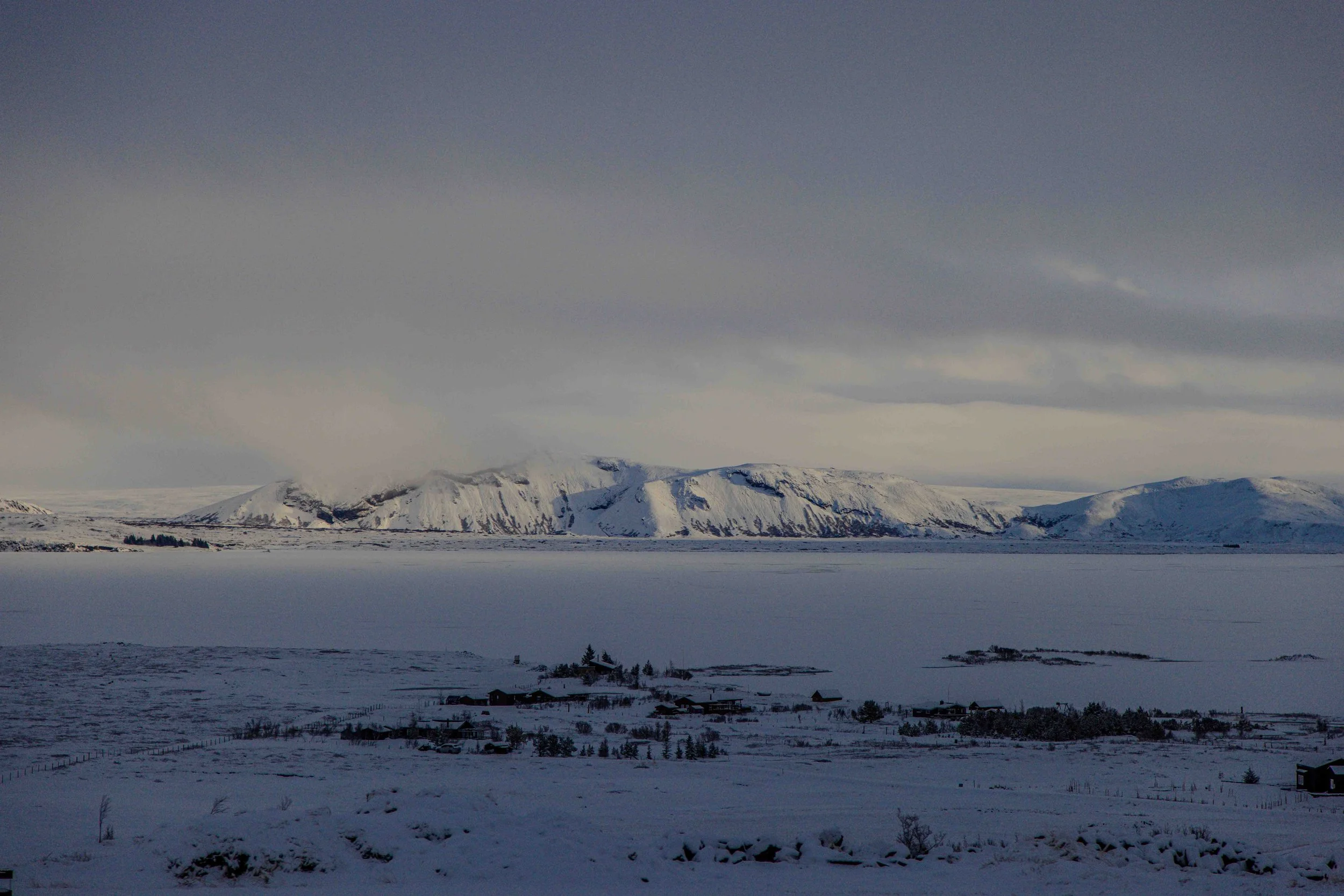 Snow-covered landscape with a frozen lake, small houses, and mountains in the background under a cloudy sky.