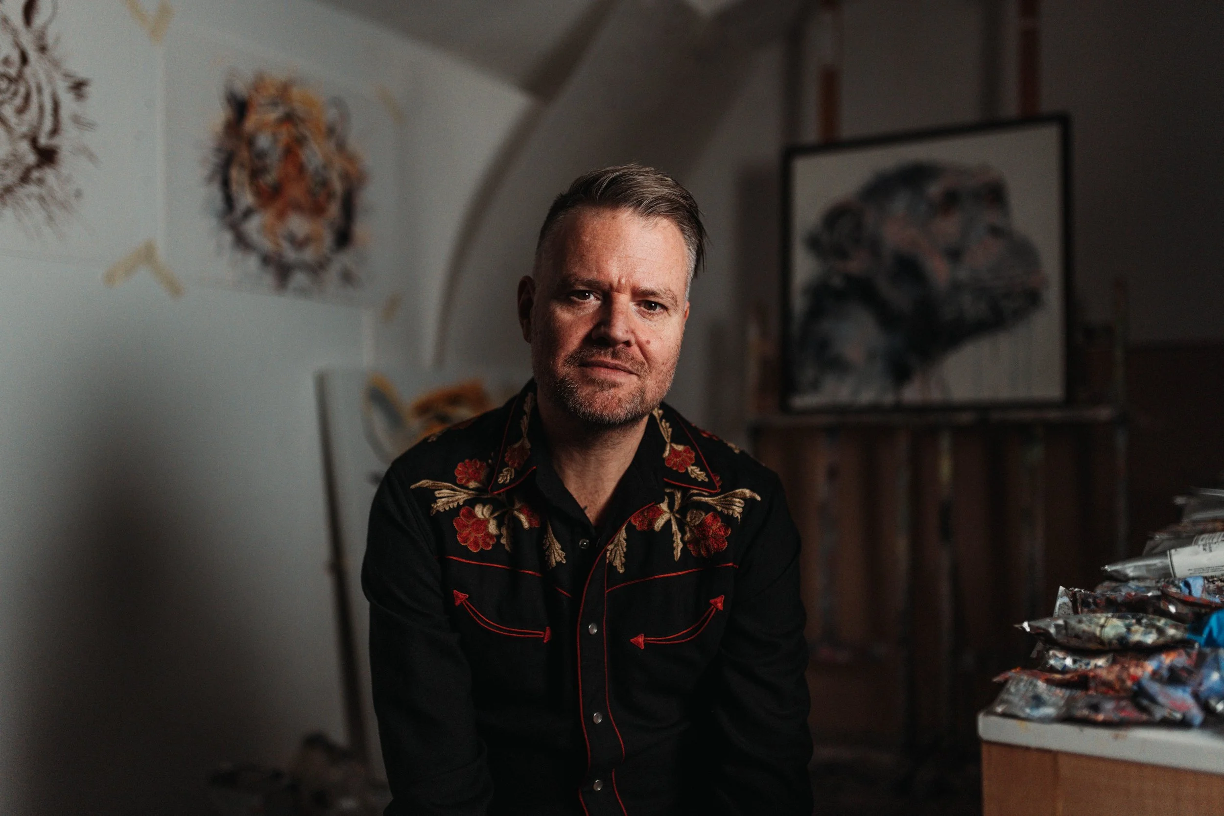 A man with short, gray hair and a beard, wearing a black shirt with red floral embroidery, sitting in an art studio with animal paintings and fabric supplies around him.