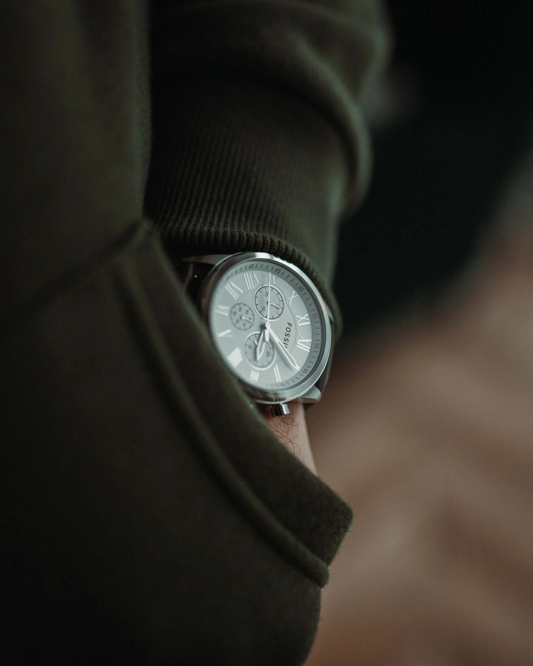 Close-up of a person's wrist wearing a Fossil watch with a gray dial and Roman numerals, partly covered by a dark green coat and a brown sleeve.
