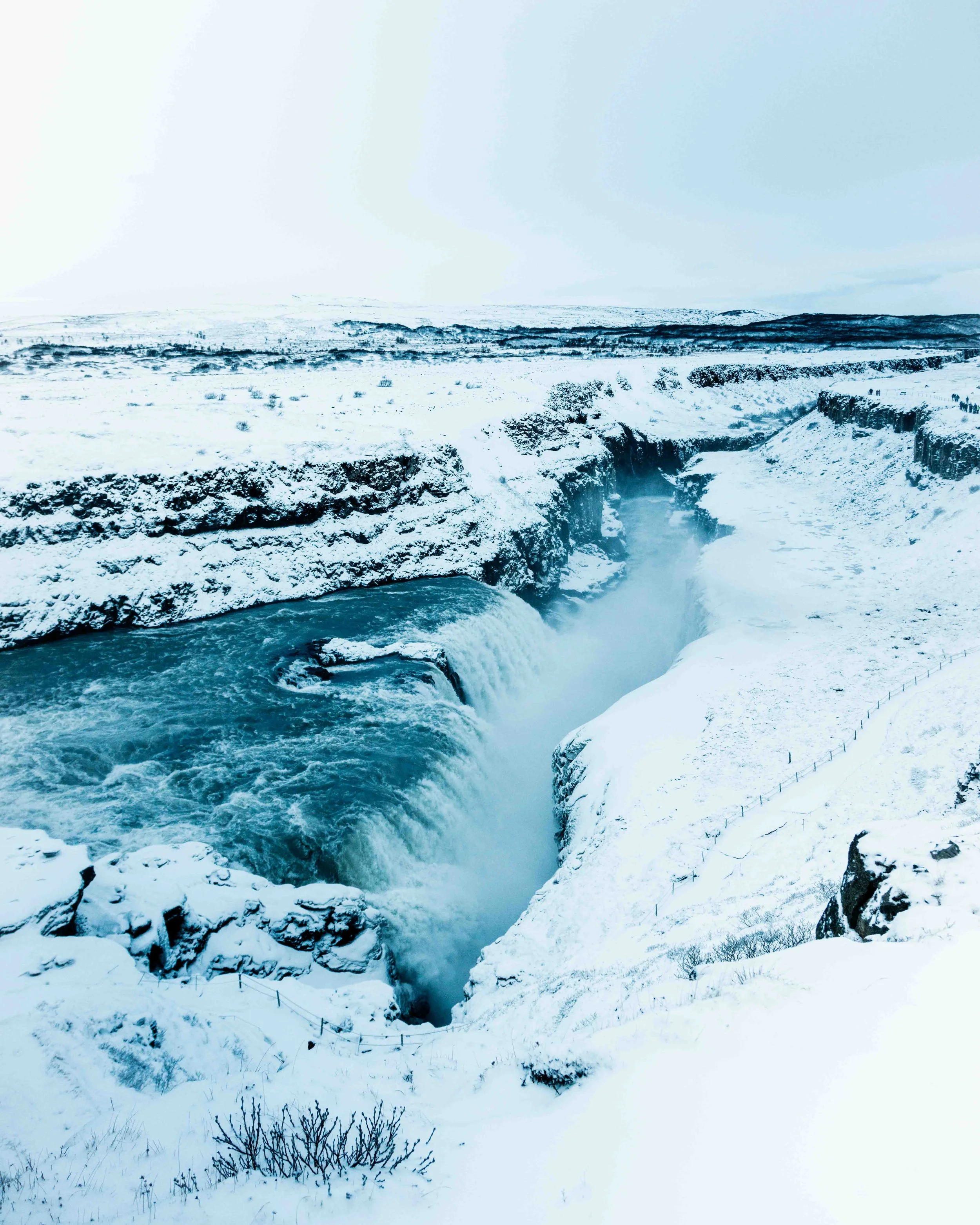 A snowy landscape featuring a waterfall flowing through a deep canyon, surrounded by snow-covered ground and sparse vegetation, under a cloudy sky.