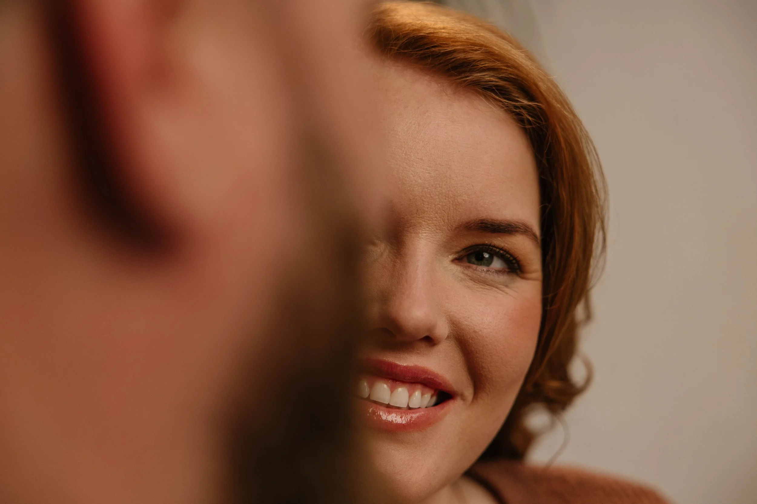 A woman with short curly red hair smiling and looking toward the camera, with part of her face blurred in the foreground.