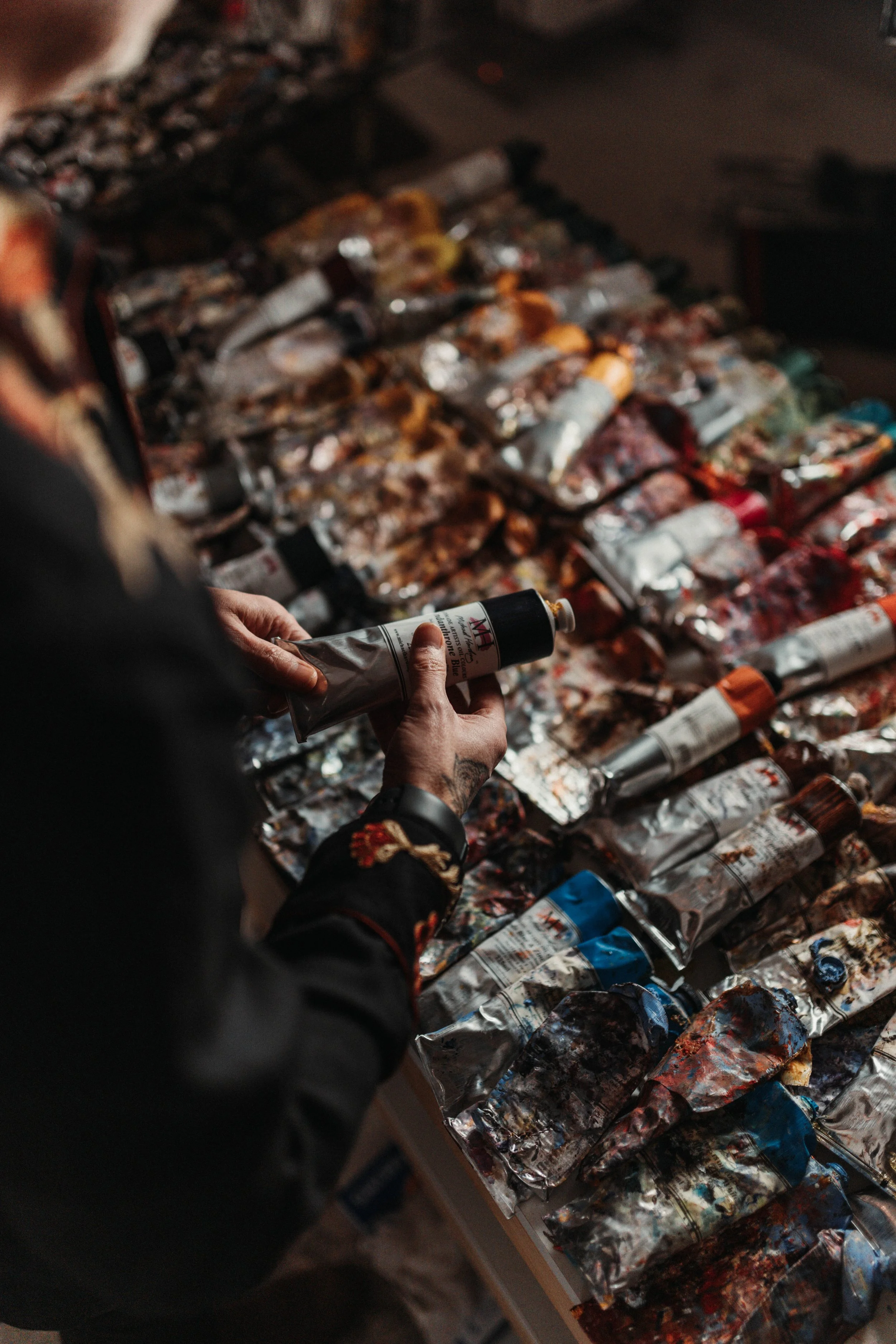 Person holding one tube of paint in front of a large table filled with colorful squeeze tubes of paint.
