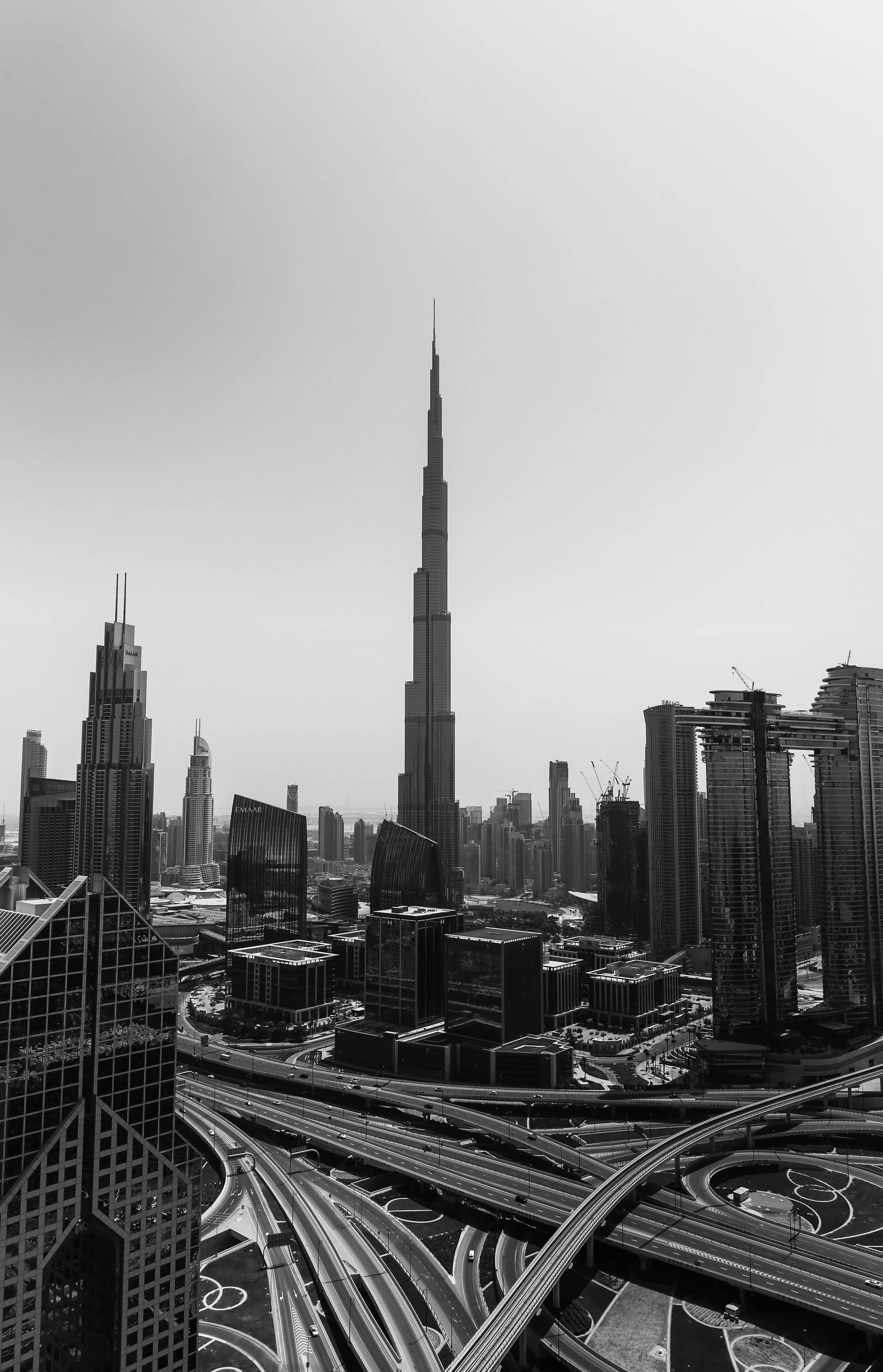 Black and white aerial view of Dubai skyline featuring the Burj Khalifa, surrounded by modern skyscrapers and busy highway interchange.