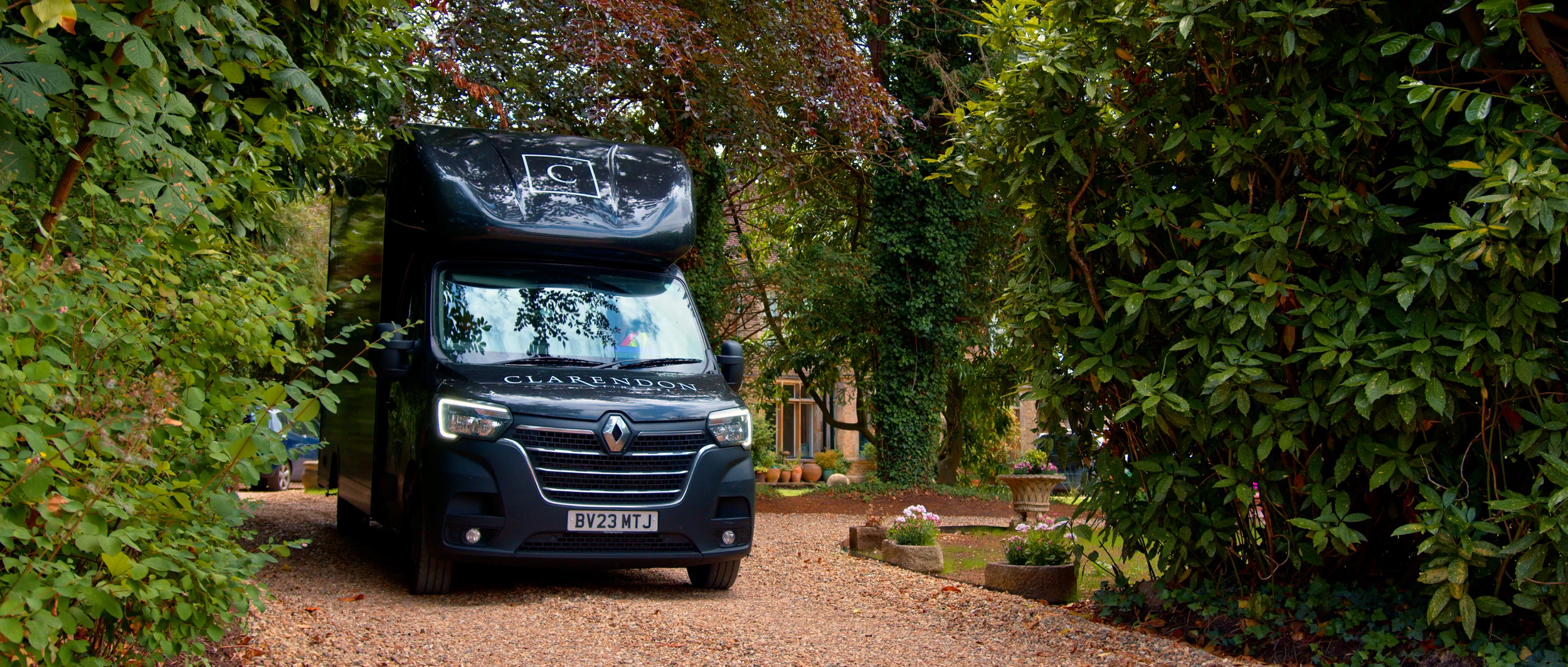 Black delivery van with the word "Clarendon" on the front and a top storage compartment, parked on a gravel driveway surrounded by green foliage and trees.