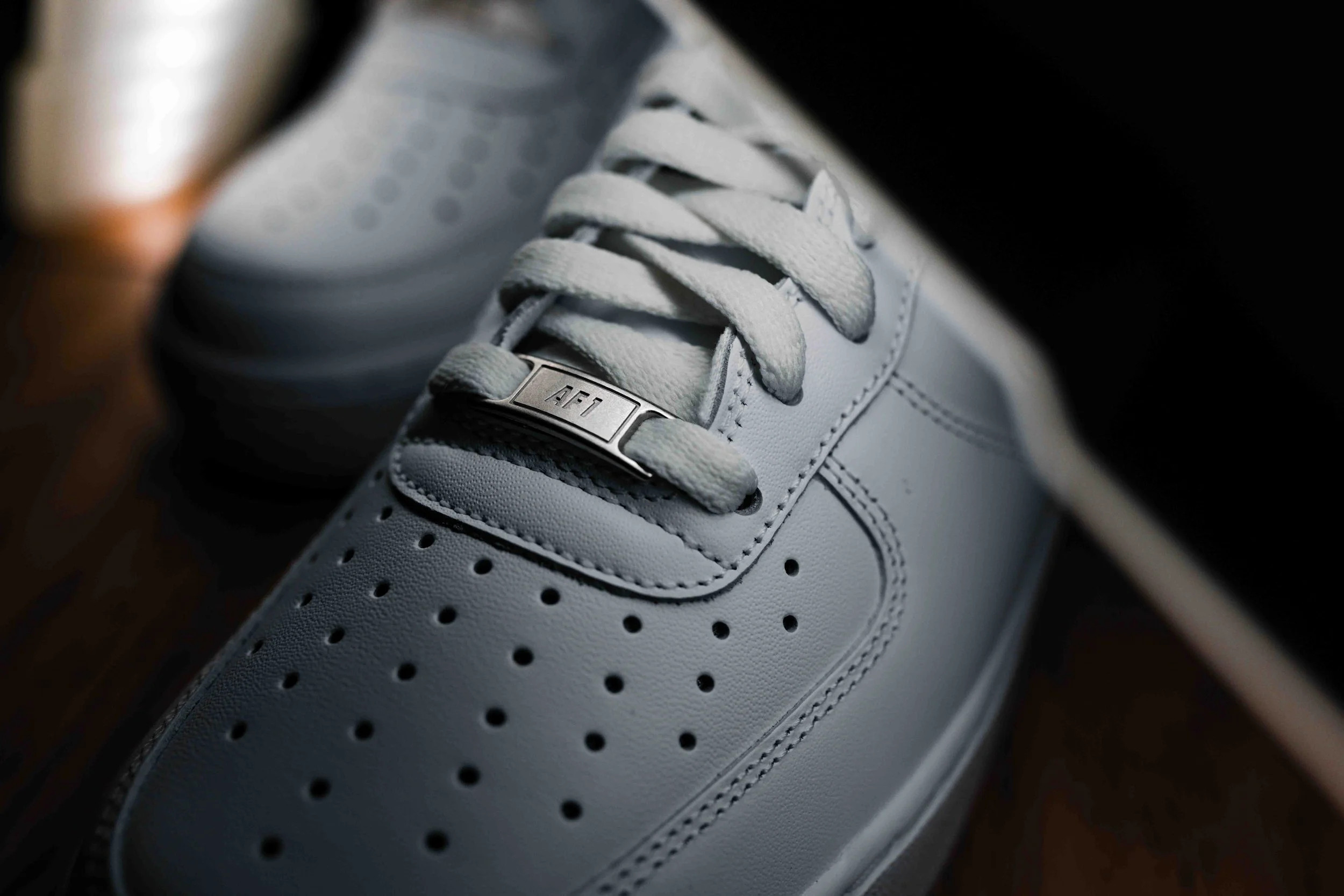 Close-up of a white athletic shoe with perforations and a metal tag on the laces reading 'AFT'.