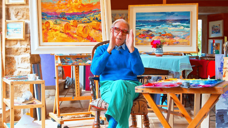 An elderly man with glasses and a blue sweater sitting in an art studio, surrounded by colorful landscape paintings and art supplies.