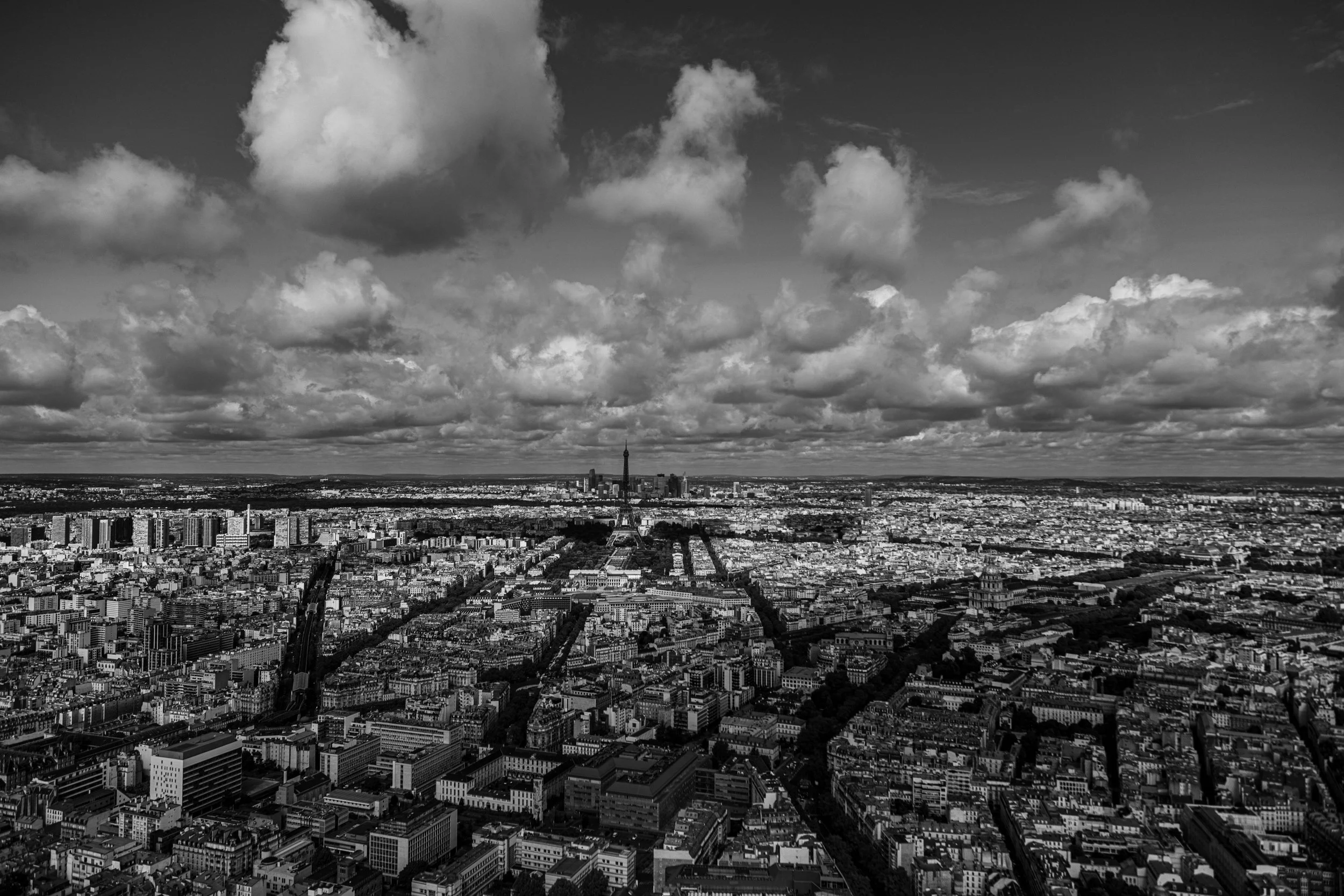 A black and white aerial view of Paris showing the Eiffel Tower in the distance and many buildings and streets below, with clouds in the sky.