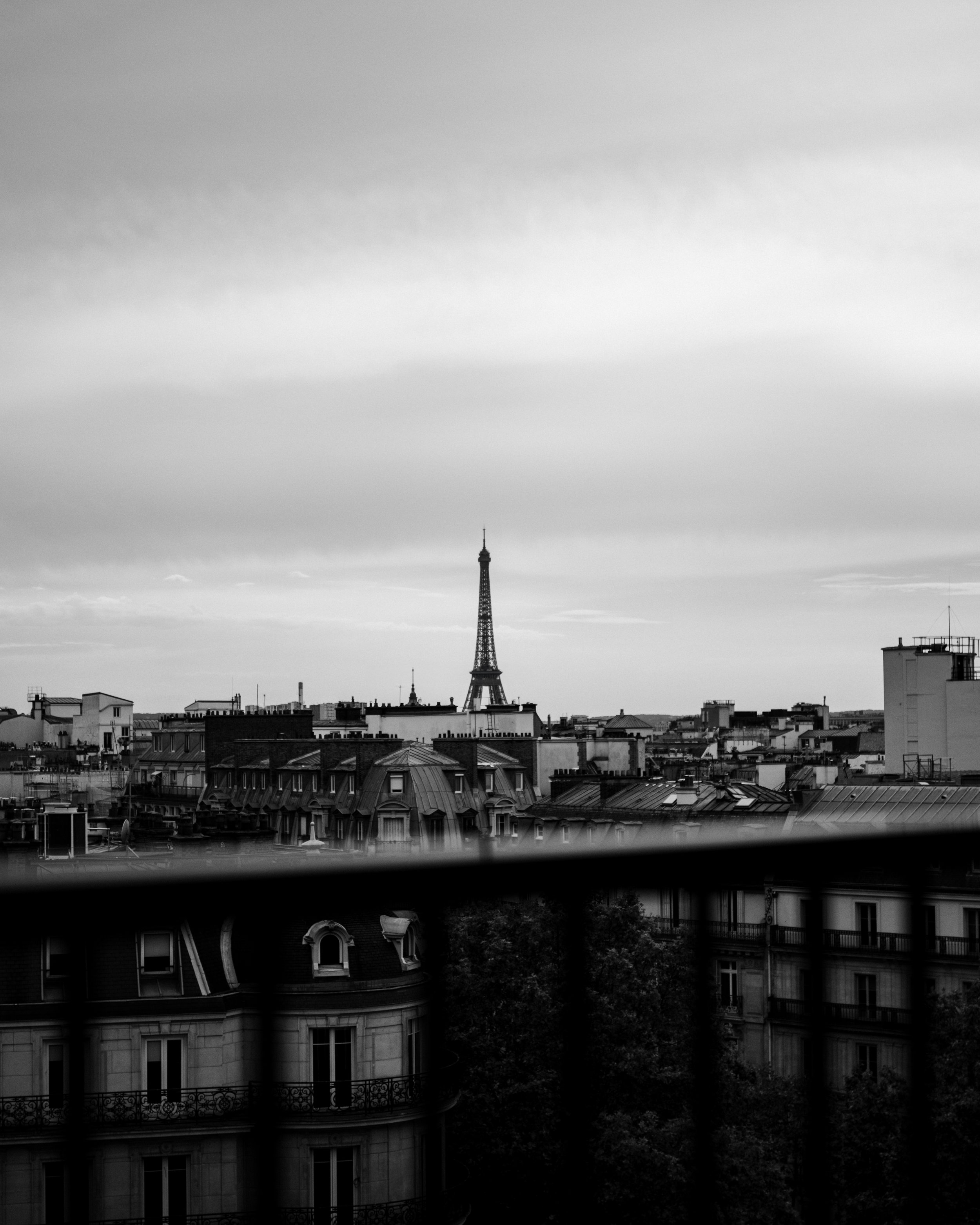 Black and white photo of the Paris cityscape with the Eiffel Tower in the distance, seen above rooftops and buildings, framed by a balcony railing in the foreground.