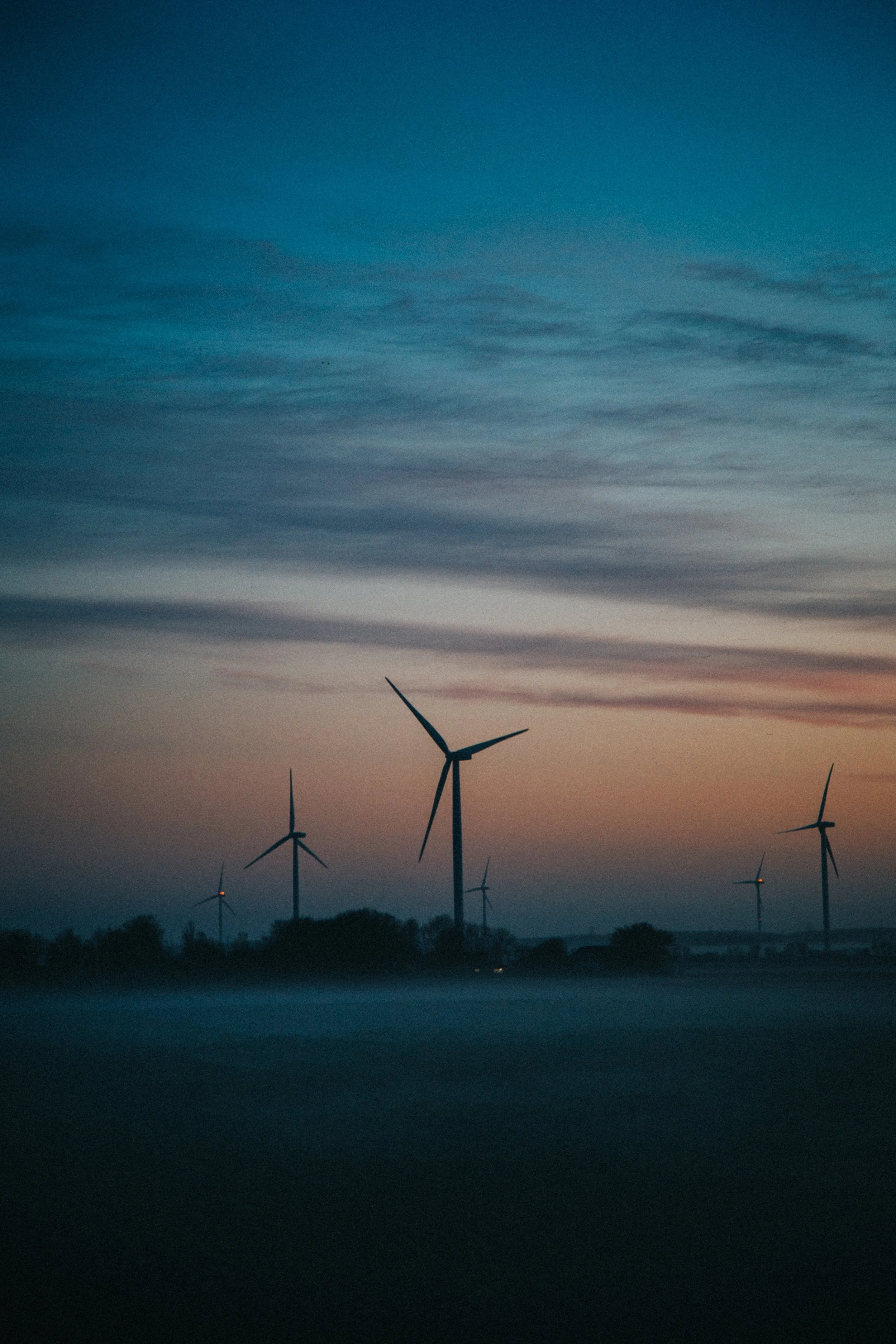 A landscape view of wind turbines at sunset or sunrise with a colorful sky and some fog or mist near the ground.