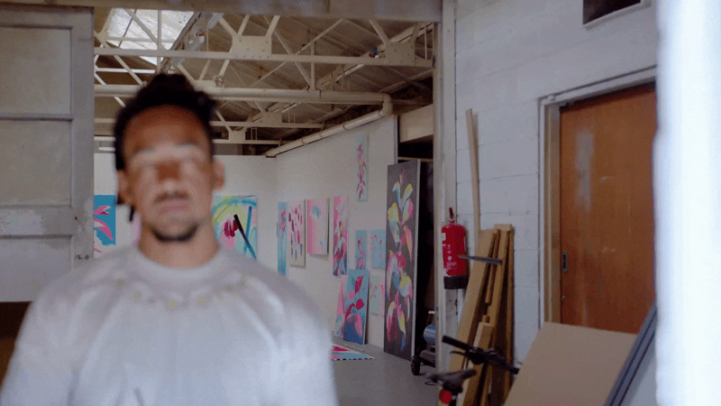Blurry person with dreadlocks standing in front of colorful abstract paintings on a gallery wall.