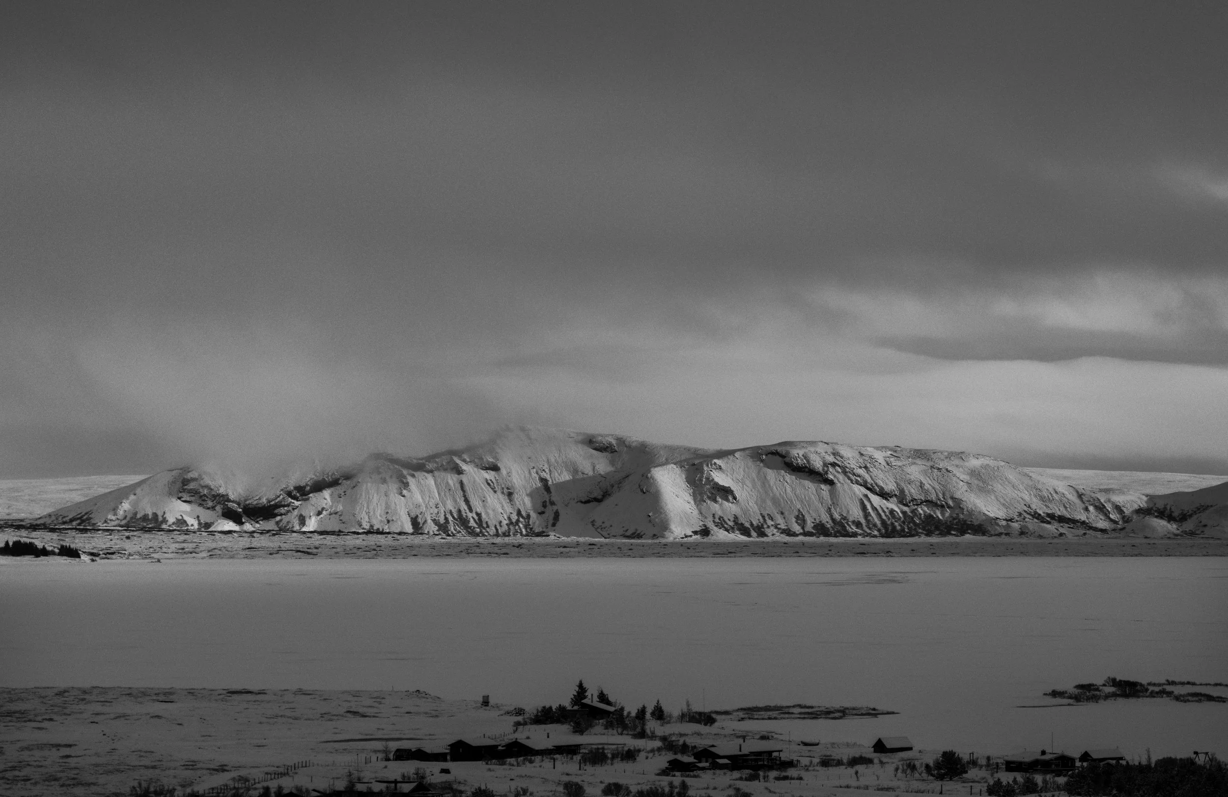 Snow-covered mountains under a cloudy sky, with a frozen landscape and small houses in the foreground.